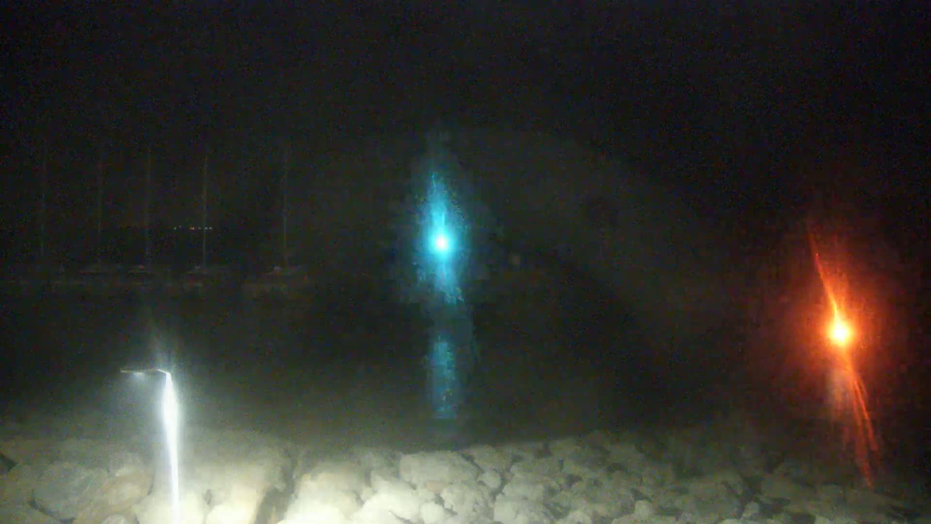 At night, three bright lights—one teal, one orange, and one white—are visible from a rocky shoreline near a dark body of water with faintly visible objects in the distance.