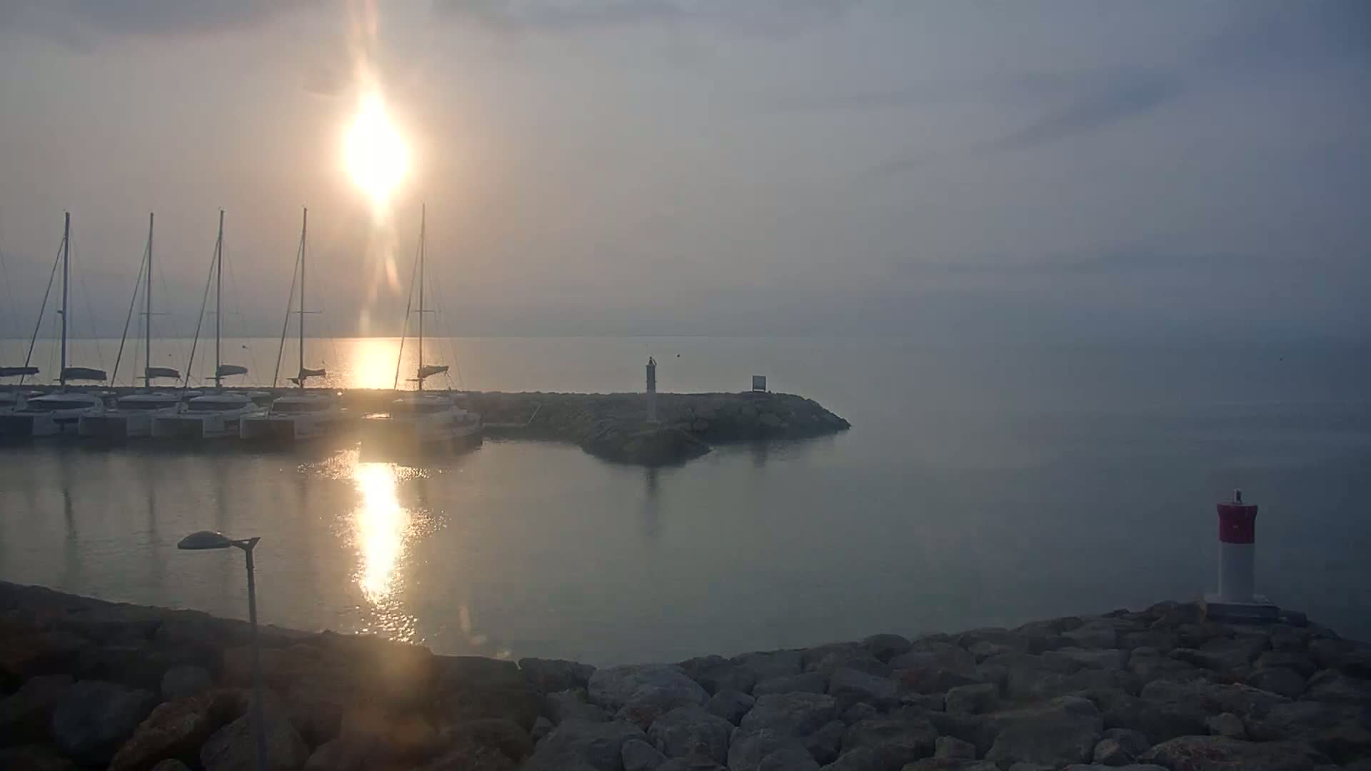 Several sailboats are docked in a calm harbor at sunrise, with a hazy atmosphere over the water.