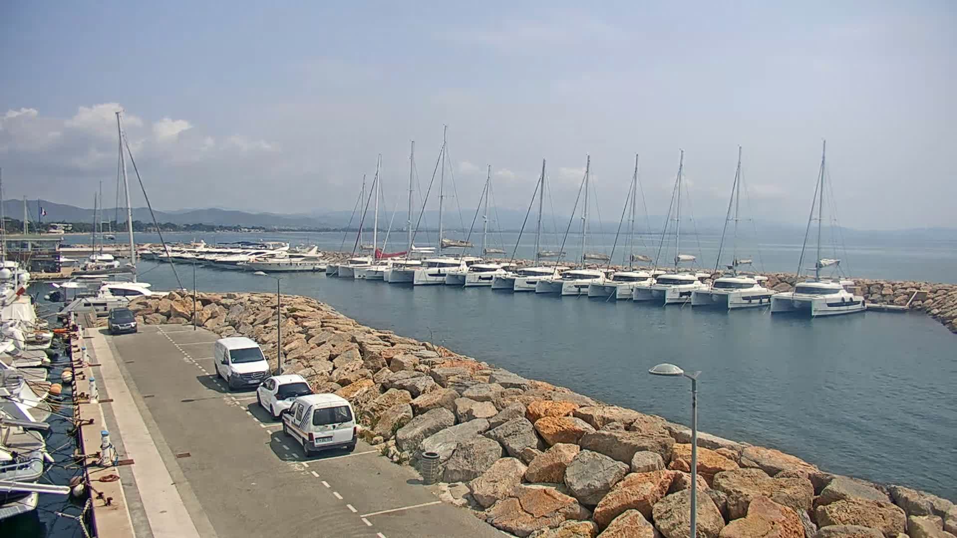 A marina with numerous sailboats and catamarans is nestled beside a rocky shore on a sunny day with a few clouds.
