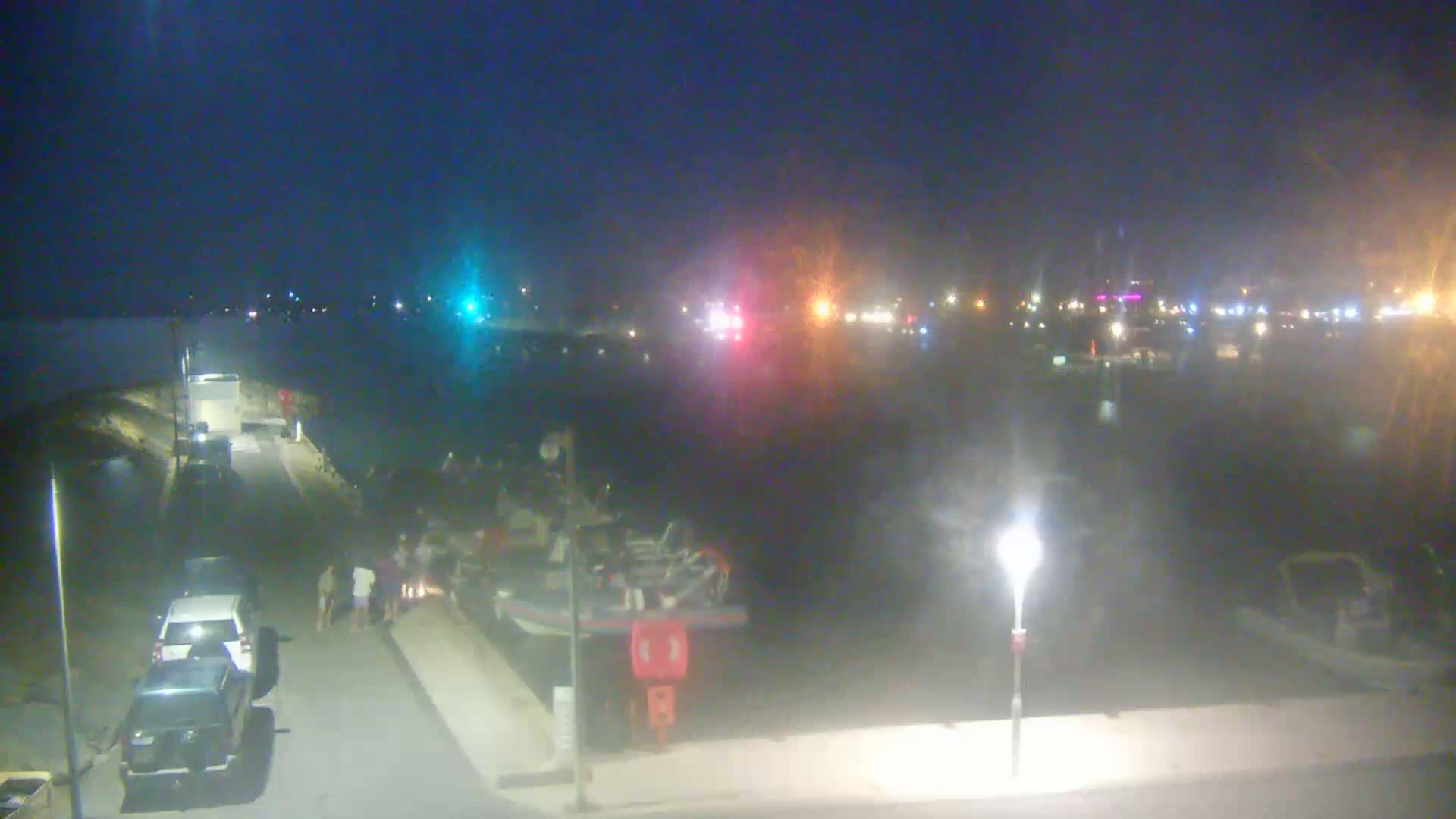 At night, a group of people stand near a boat docked at a waterfront, with vehicles parked nearby and lights illuminating a calm body of water in the background.