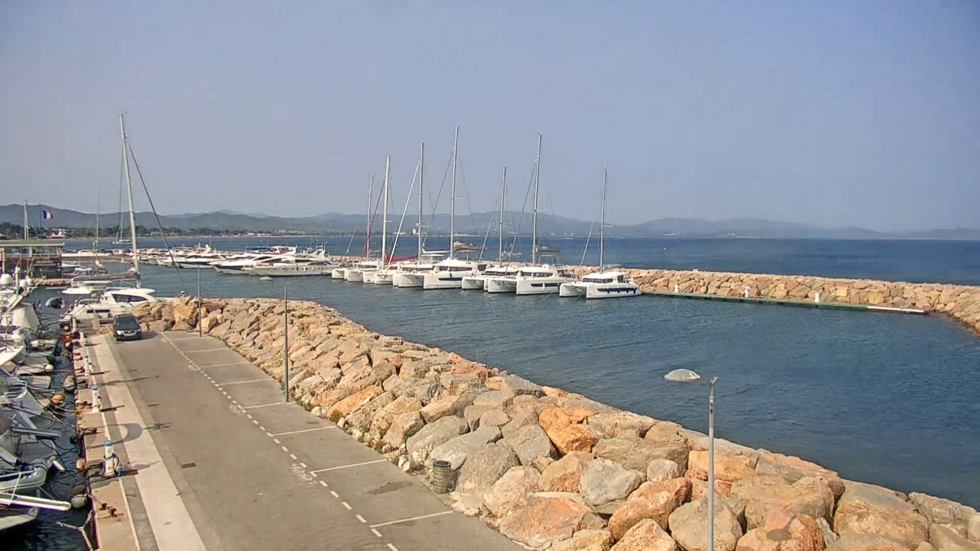 A marina with many boats, including several catamarans, is situated behind a rock wall on a calm, sunny day.