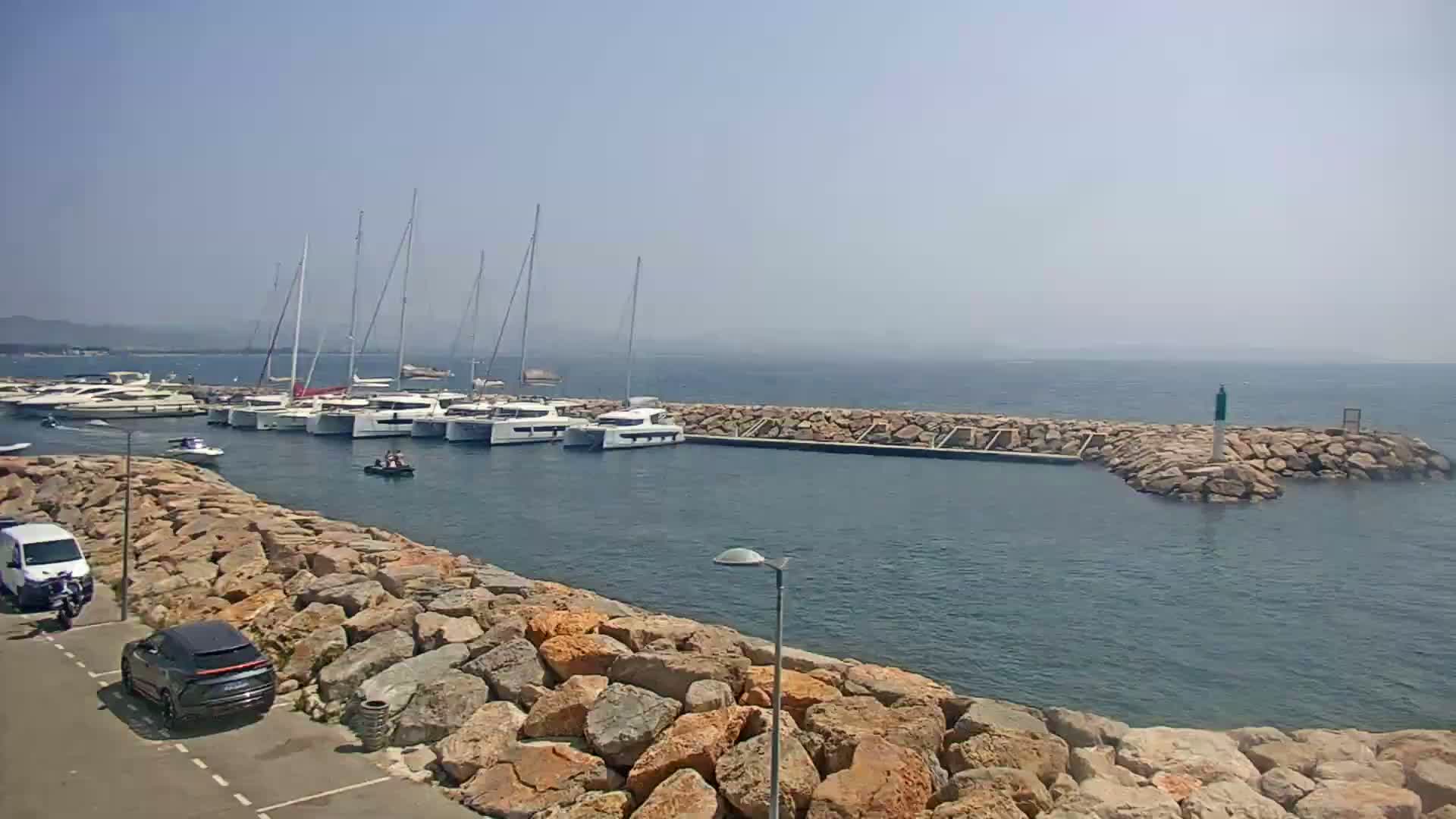 A marina filled with sailboats and motor yachts is partially enclosed by a rocky breakwater on a clear, sunny day.