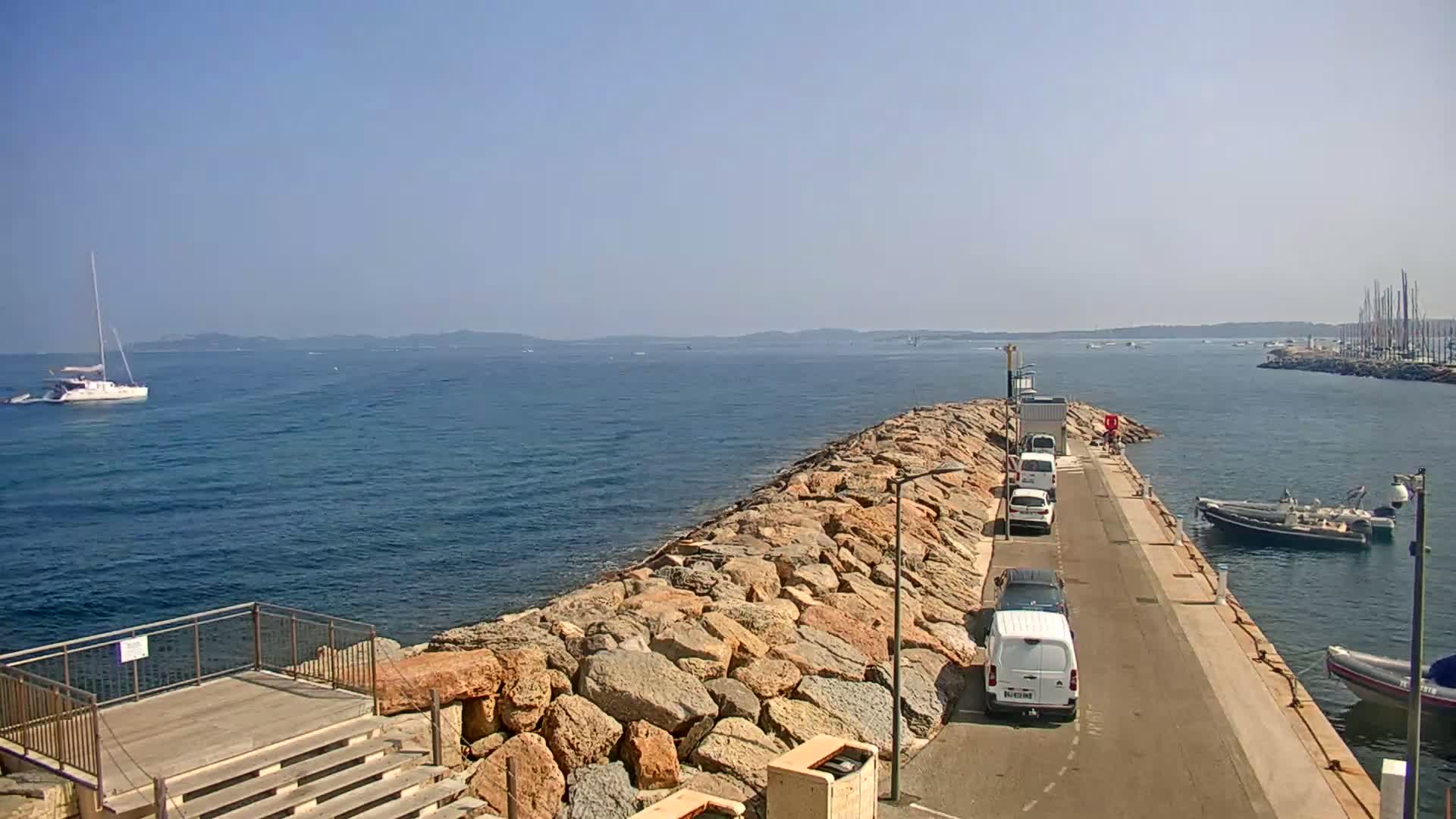 A rocky breakwater extends into a calm blue sea, where several boats are moored, under a clear, sunny sky.