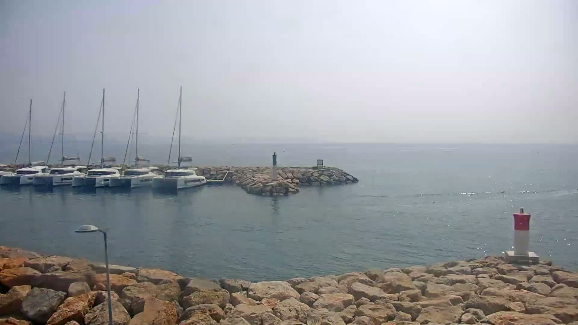 Several sailboats are docked in a harbor on a hazy day, with a small rocky island and a navigational marker visible in the water.
