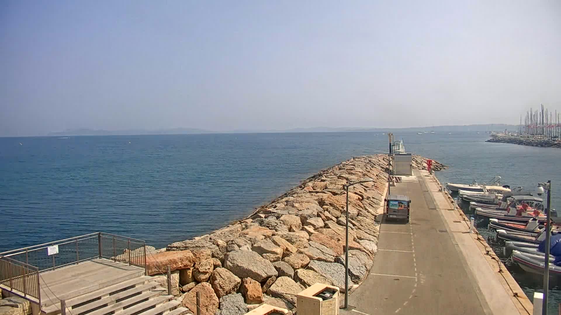 A rocky breakwater extends from a paved road lined with parked boats into a calm, blue sea under a clear sky.
