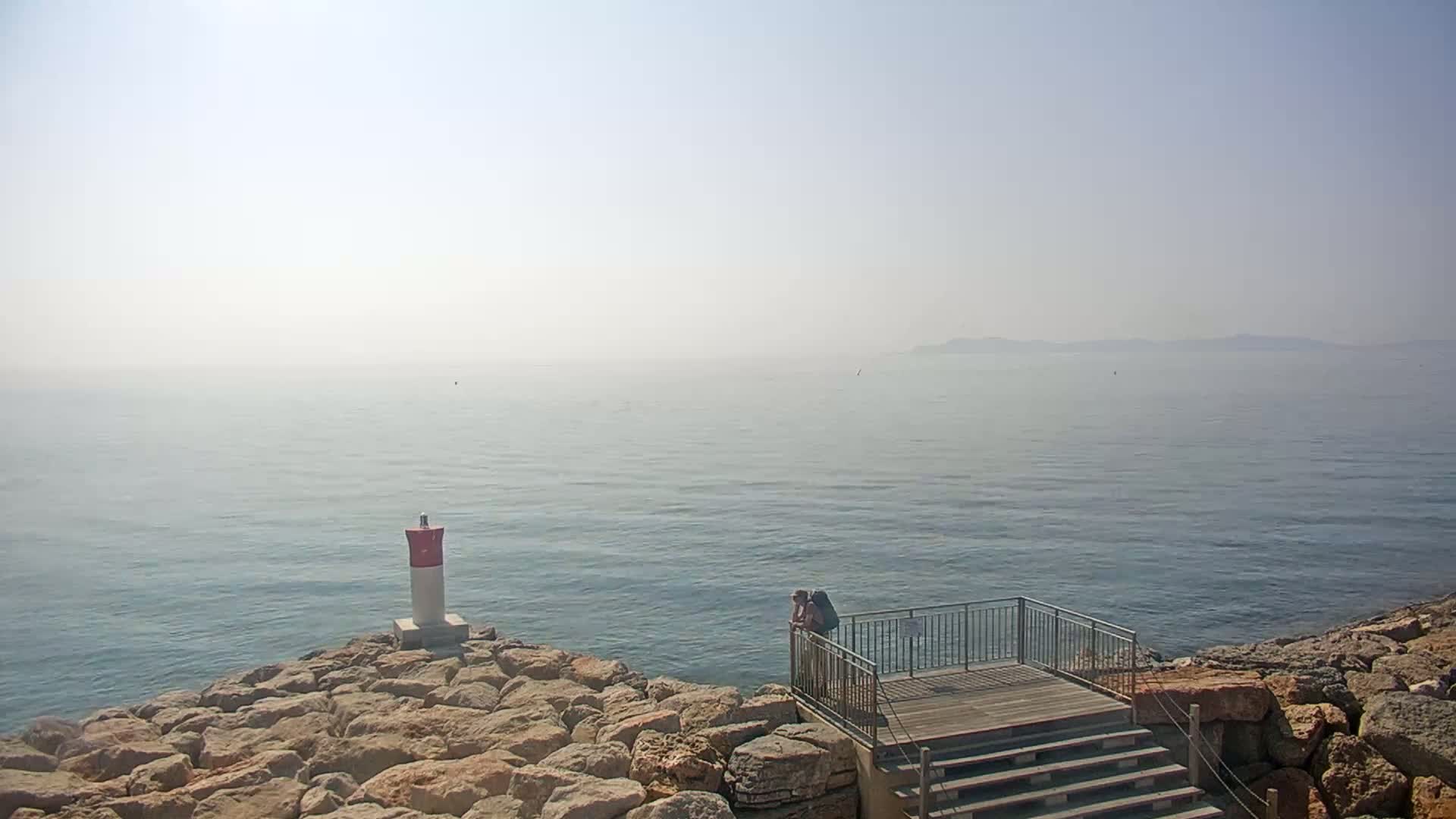 A person stands on a wooden platform with stairs overlooking a hazy, calm sea with a small, red and white marker on a rocky shoreline.