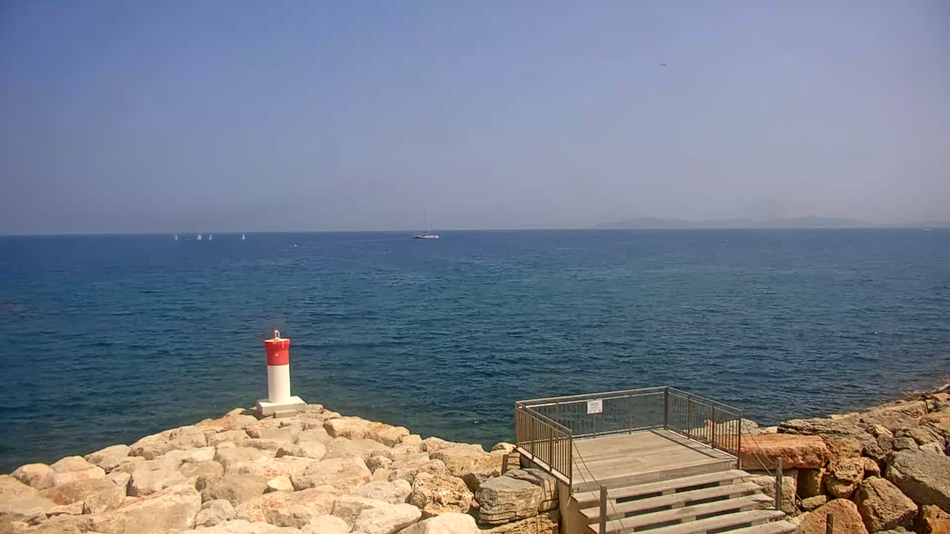 A rocky shoreline with a small wooden platform and stairs overlooks a calm, blue sea on a clear, sunny day, with a few sailboats visible in the distance.
