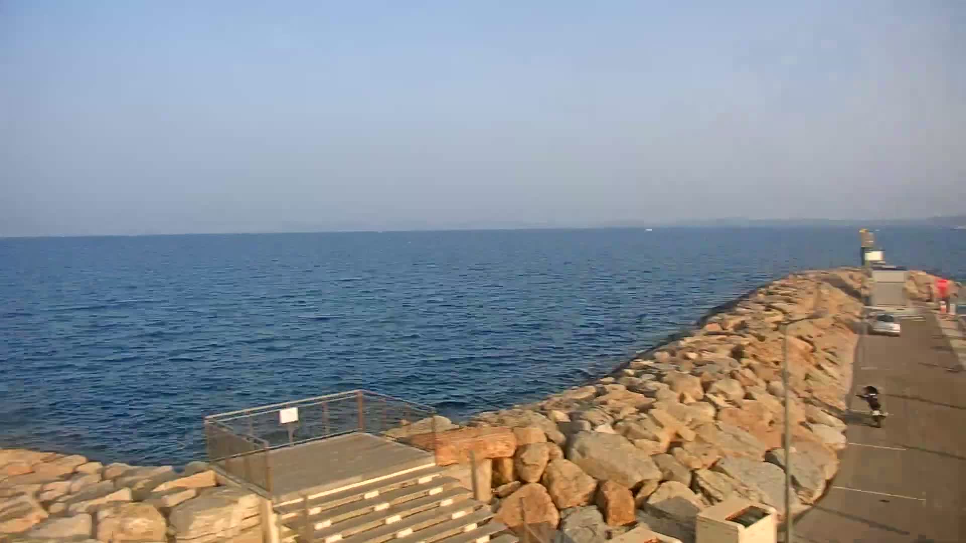 A rocky breakwater extends into a calm, blue sea under a clear sky.