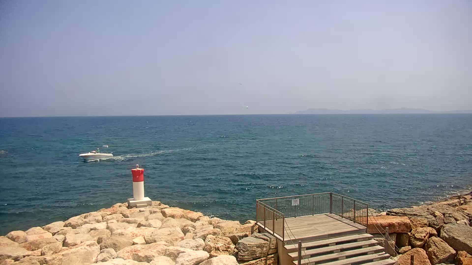A small motorboat travels on a calm, blue sea near a rocky shore with a small wooden platform and a red and white marker buoy.