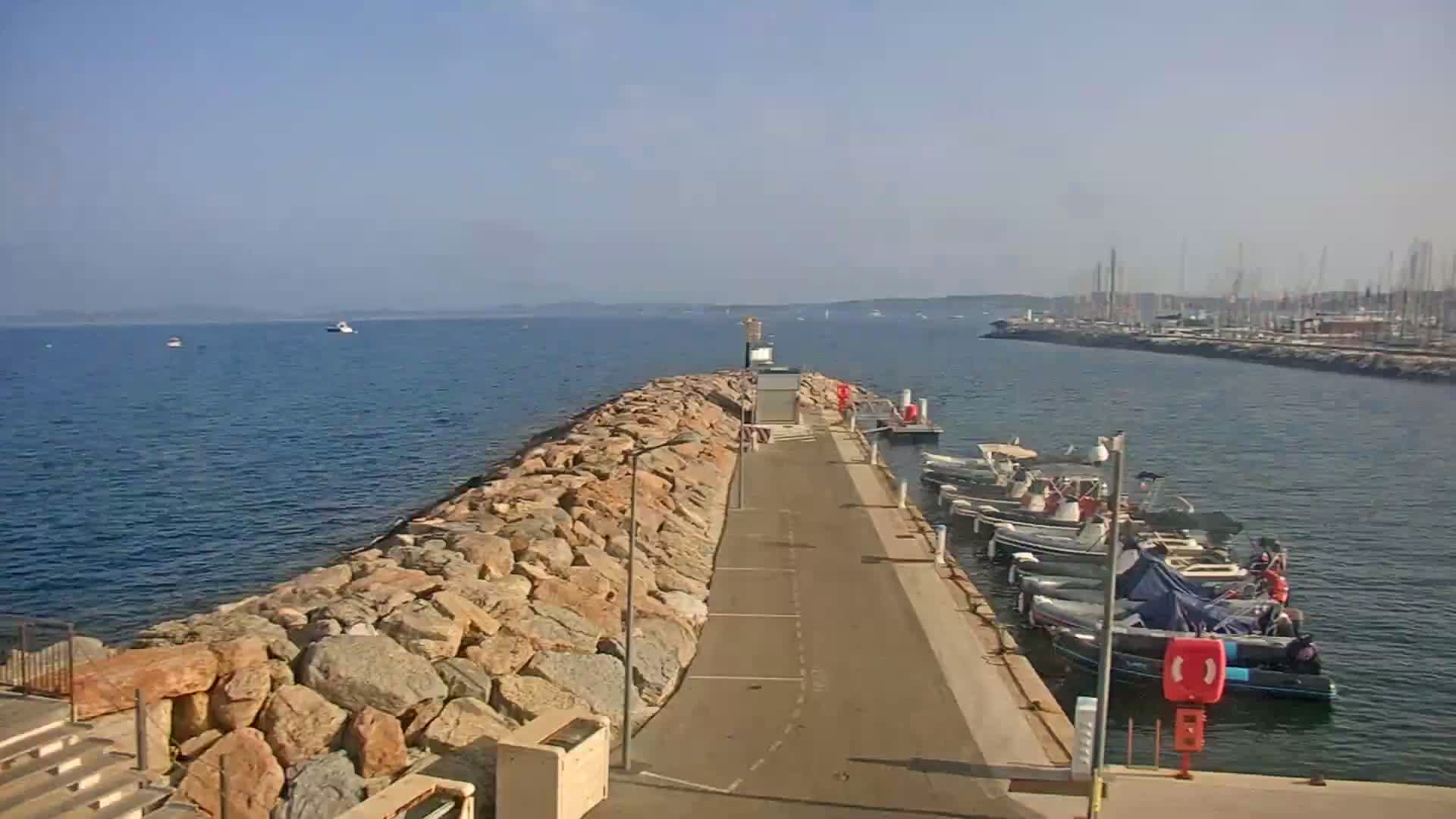 A rocky breakwater extends from a paved walkway lined with docked motorboats into a calm, blue sea on a sunny day.