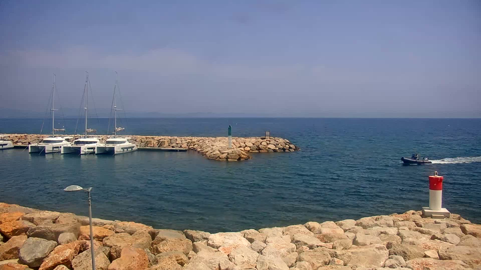 Several sailboats are moored in a calm, sunny harbor protected by a rock jetty, with a small motorboat passing by.