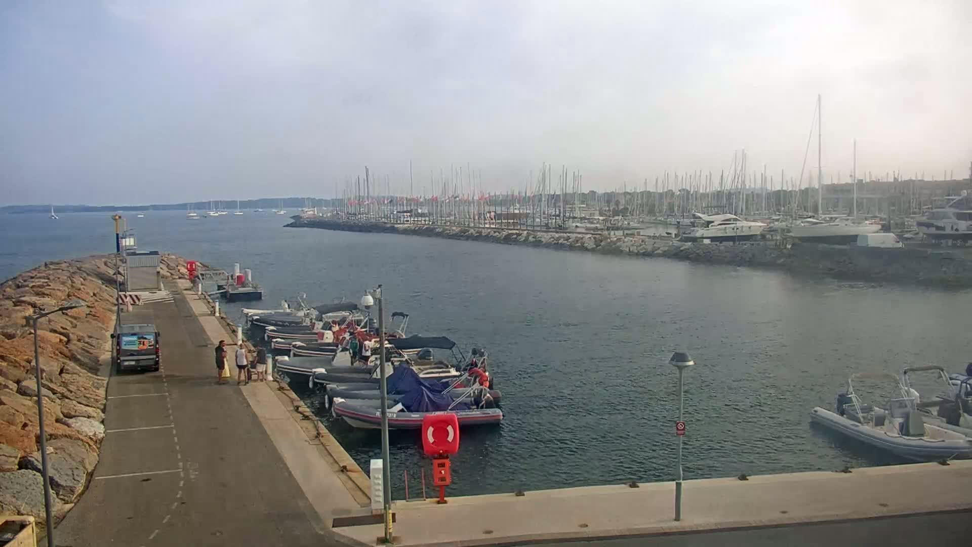 A harbor with numerous boats and yachts moored, viewed from a stone jetty on a calm, hazy day.