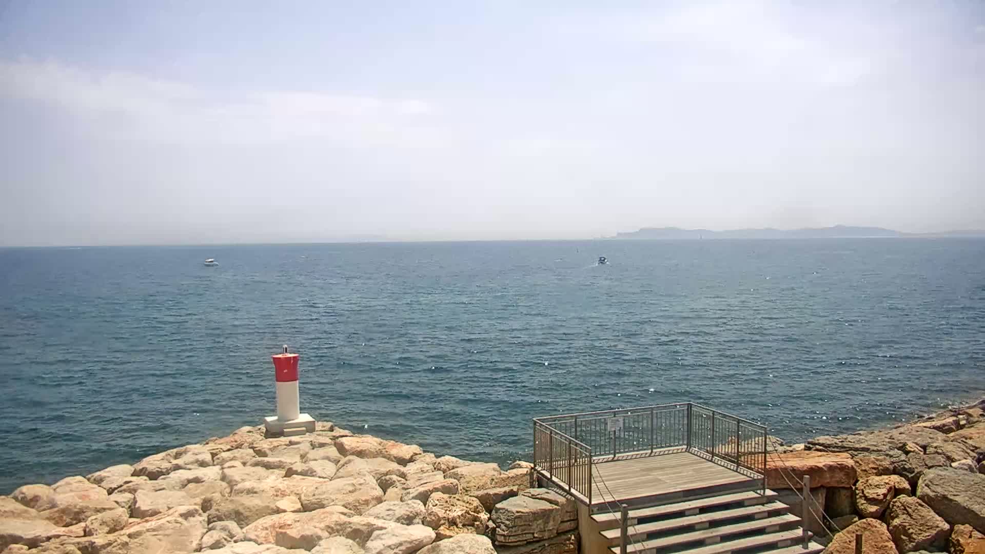 A rocky shoreline with a small viewing platform overlooks a calm sea under a clear sky, with a distant landmass visible on the horizon.