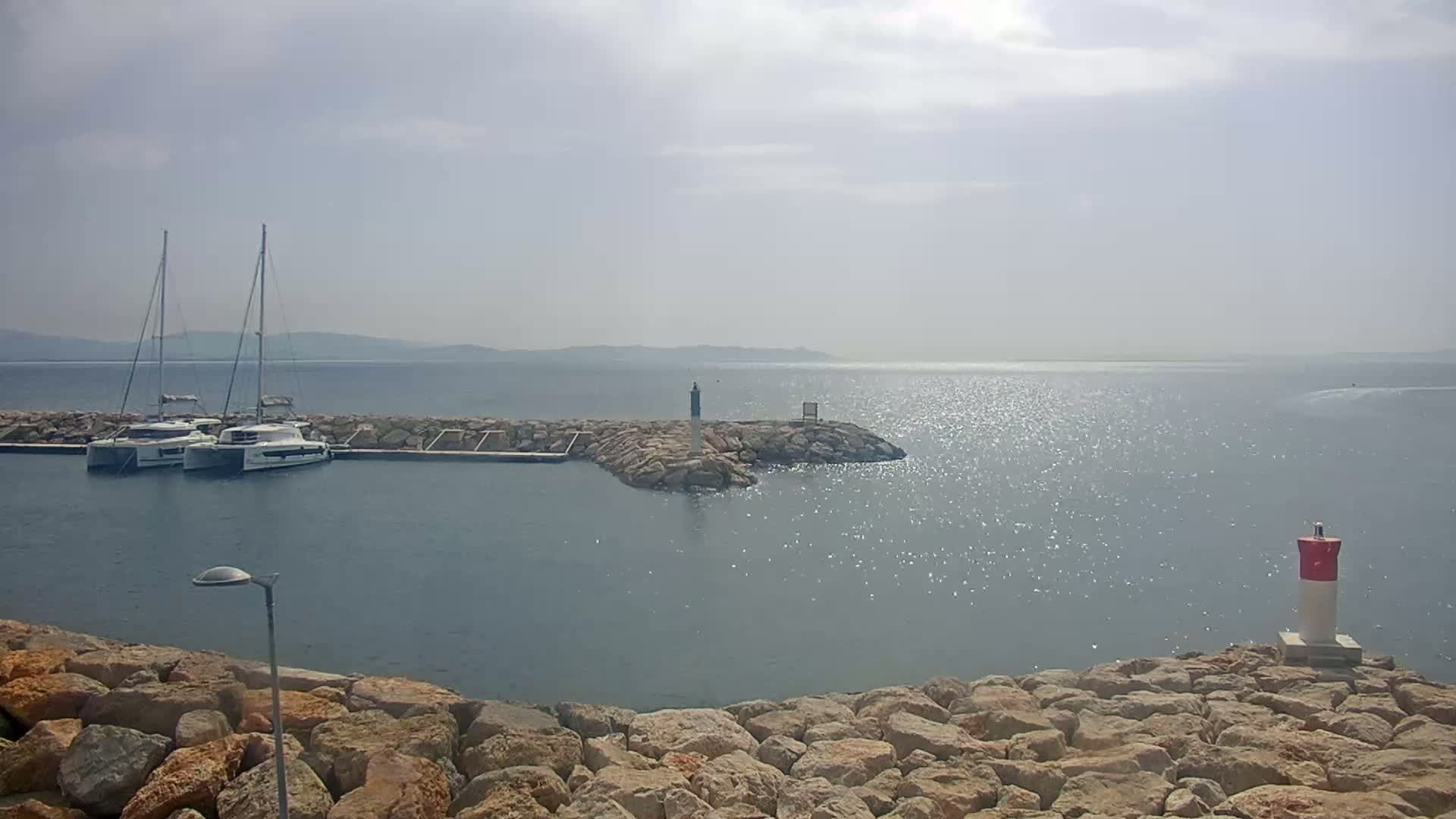 Two sailboats are docked in a calm harbor on a partly sunny day, with a rocky shoreline and distant hills visible across the sparkling water.