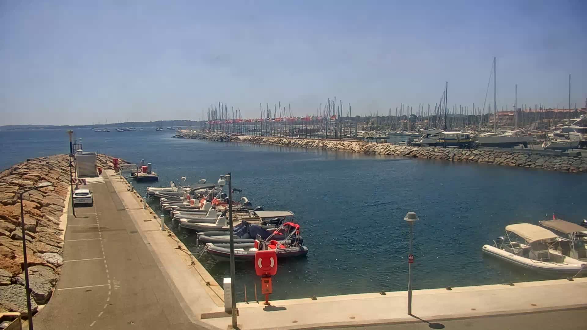 A sunny day at a marina shows many boats docked along a stone wall, with additional small motorboats moored at a nearby pier.