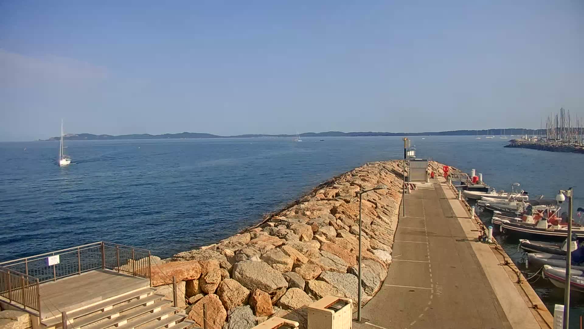 A rocky breakwater extends into a calm blue sea, with several boats moored at its end and a sailboat in the distance under a clear sky.