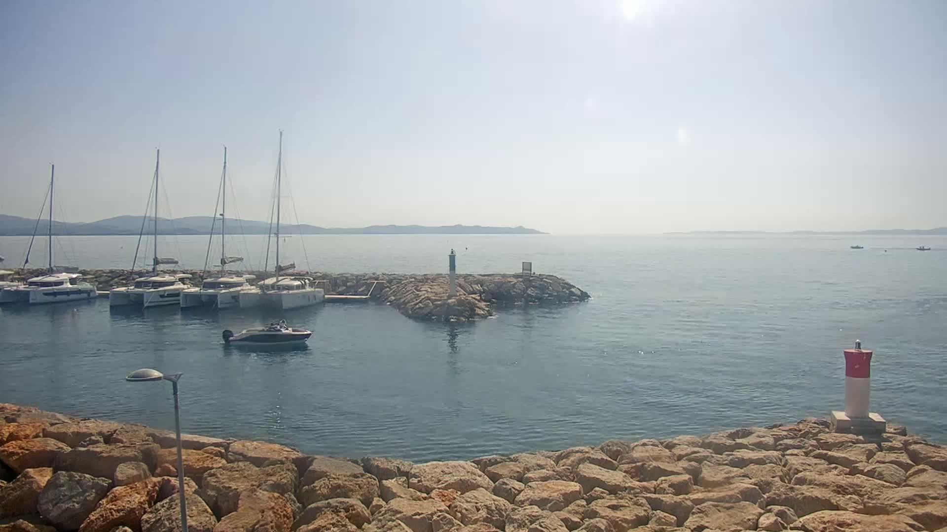 Several sailboats are moored in a calm, sunny harbor, with a rocky breakwater in the foreground and distant hills visible across the water.