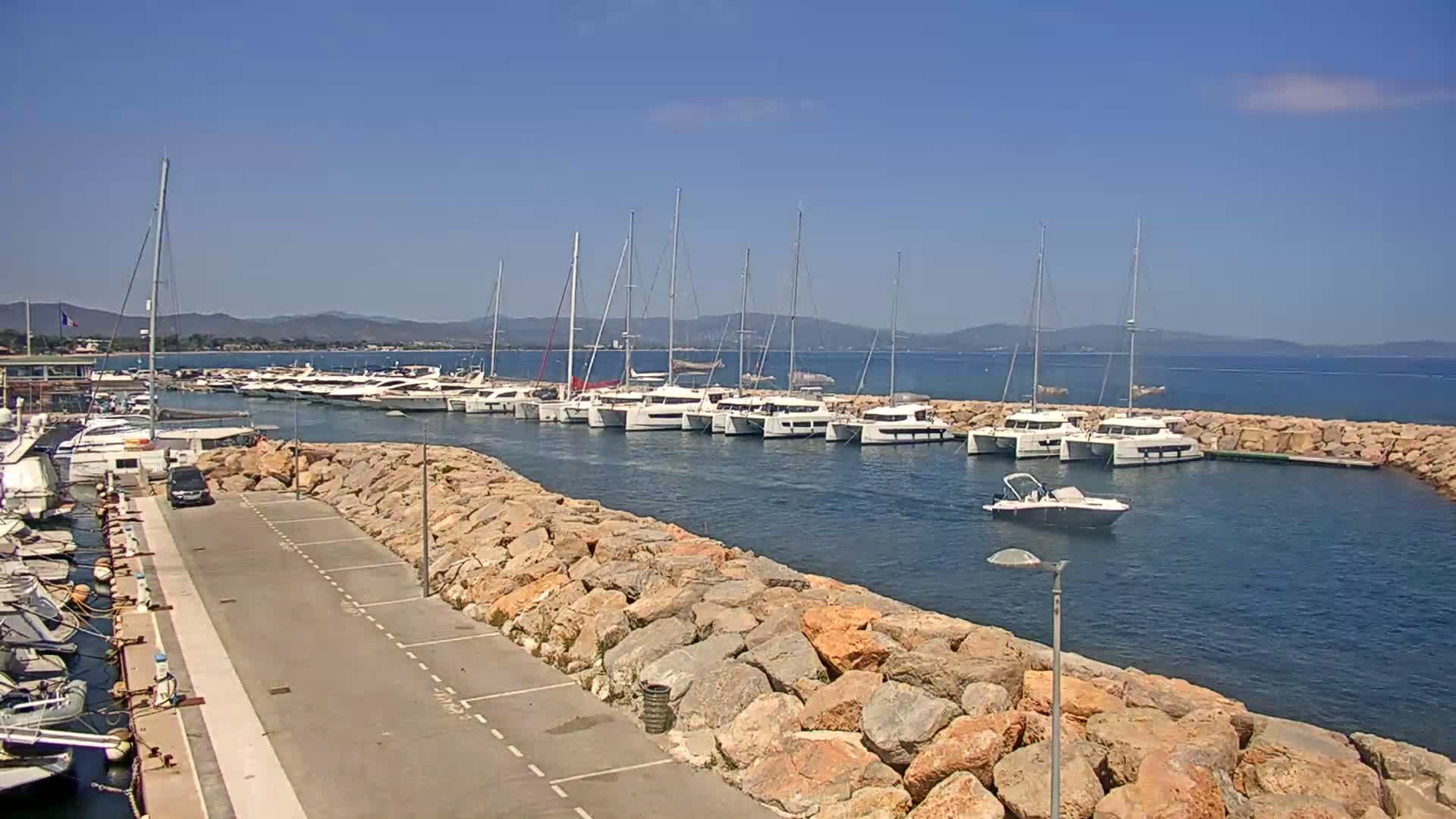 A marina filled with numerous sailboats and motorboats is situated along a rocky shoreline on a sunny day with calm waters.