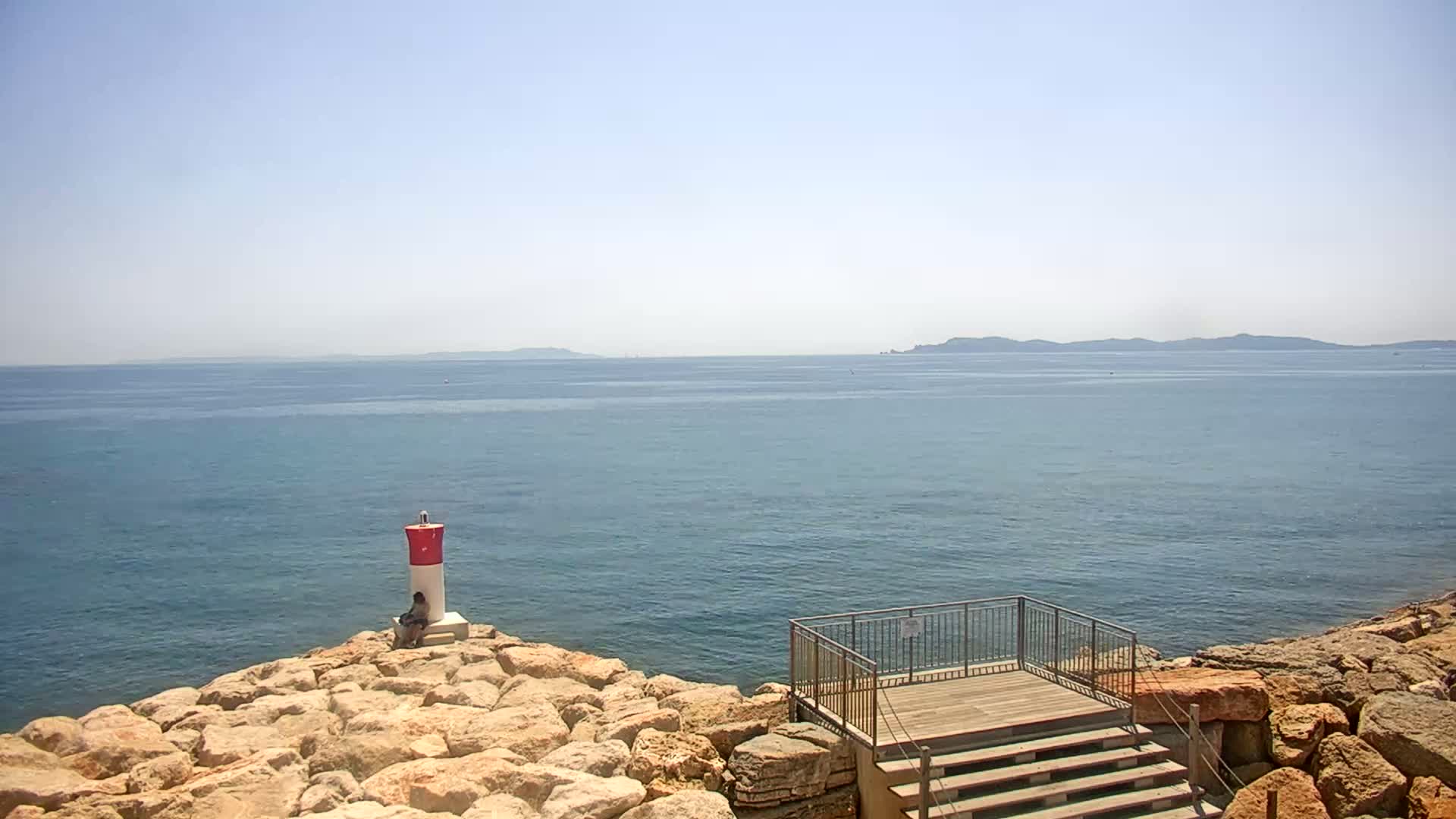 A rocky shoreline overlooks a calm, blue sea under a clear sky, featuring a small lighthouse and a wooden viewing platform with stairs.