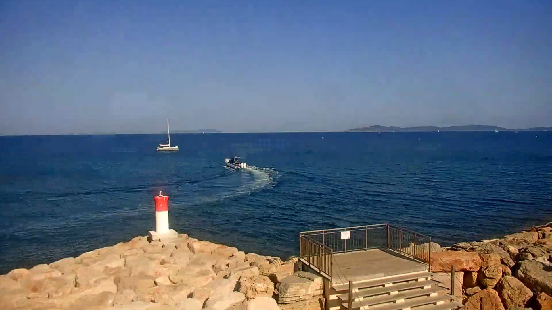 A rocky shoreline with a small viewing platform overlooks a calm blue sea where a sailboat and motorboat are visible, under a clear blue sky.