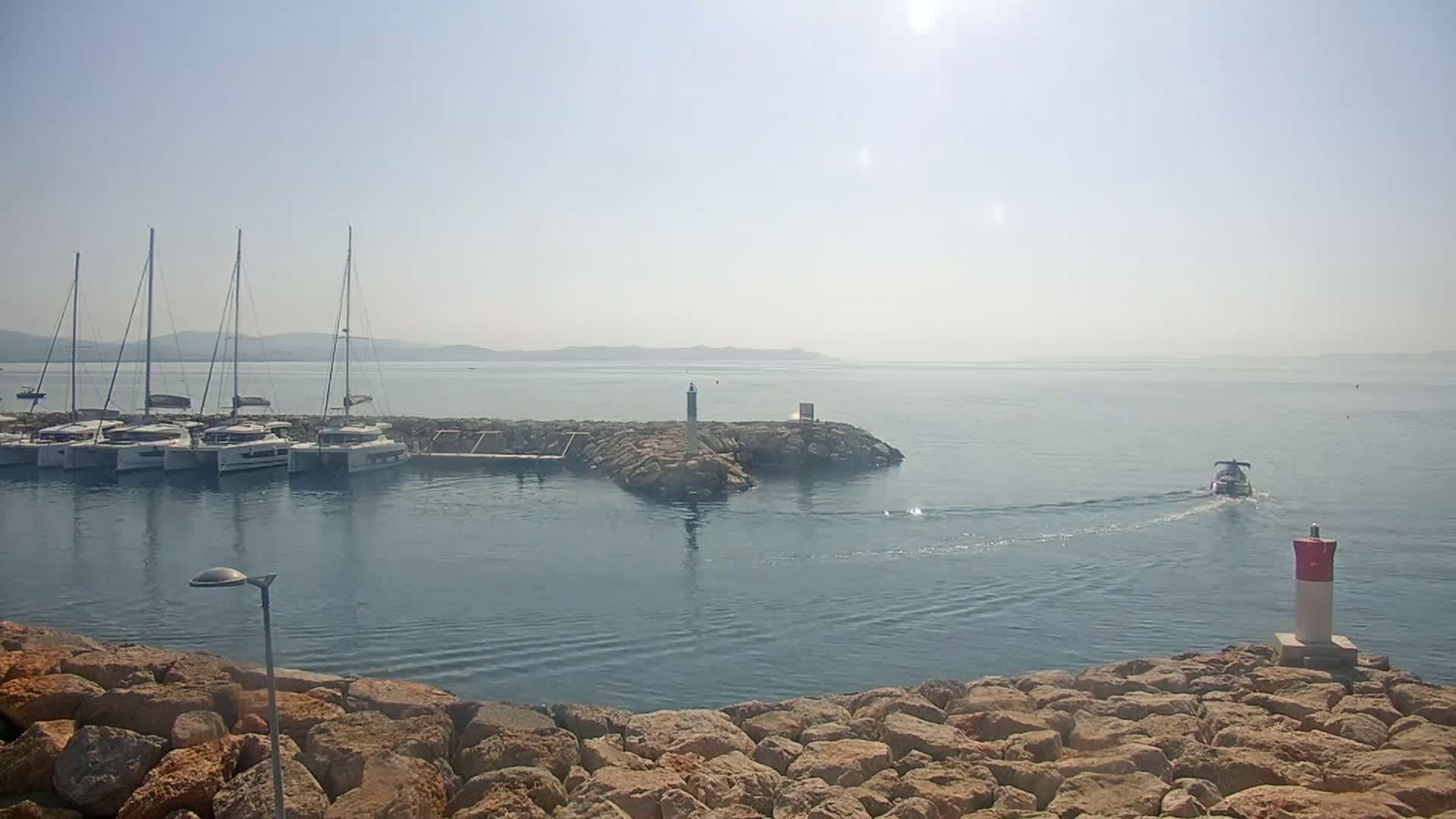 Several sailboats are moored in a harbor on a sunny day with hazy conditions in the distance, and a small motorboat is traveling away from the harbor.