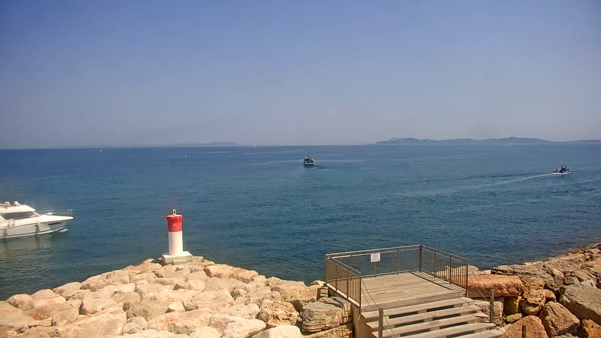 A rocky shoreline with a small wooden platform and steps overlooks a calm blue sea with several boats and distant islands under a clear sky.