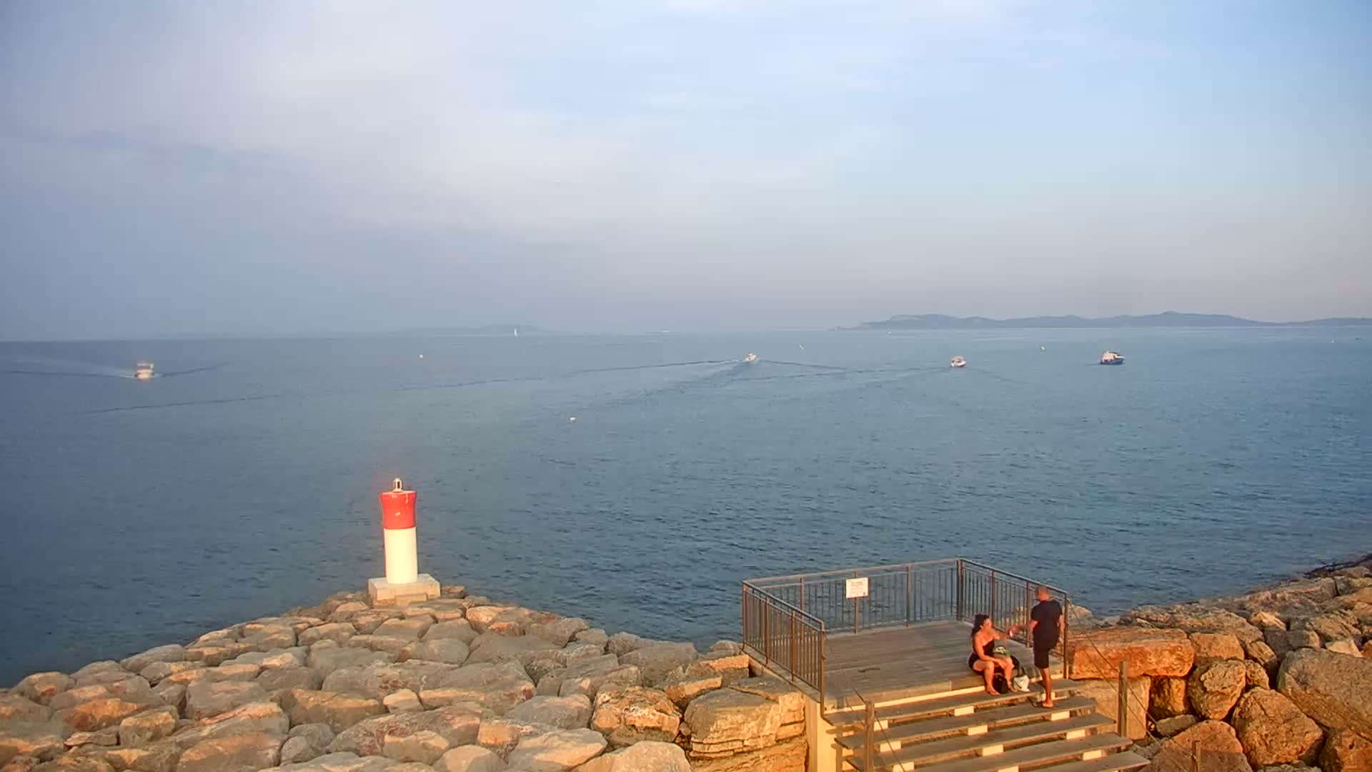 A rocky shoreline with a small viewing platform overlooks a calm sea with several boats and a distant landmass under a mostly clear sky.