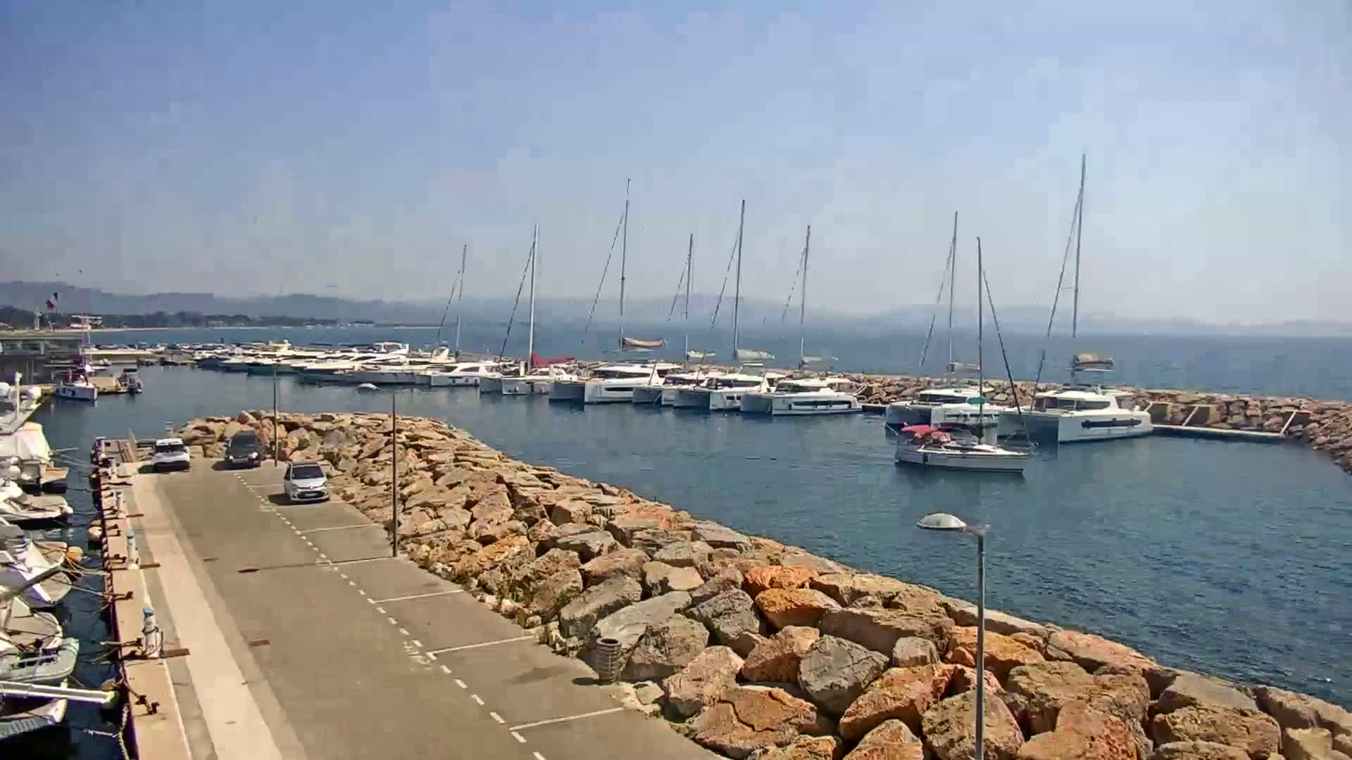 A marina filled with sailboats and motorboats is situated on a calm, sunny day next to a rocky breakwater with a paved parking lot.