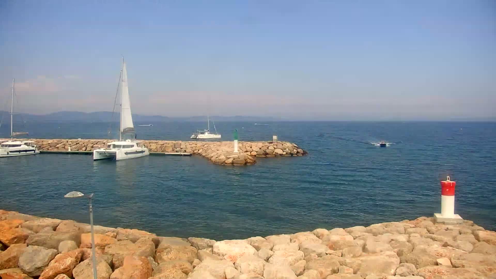 Several sailboats are moored in a calm harbor, partially enclosed by a rocky breakwater on a clear, sunny day.