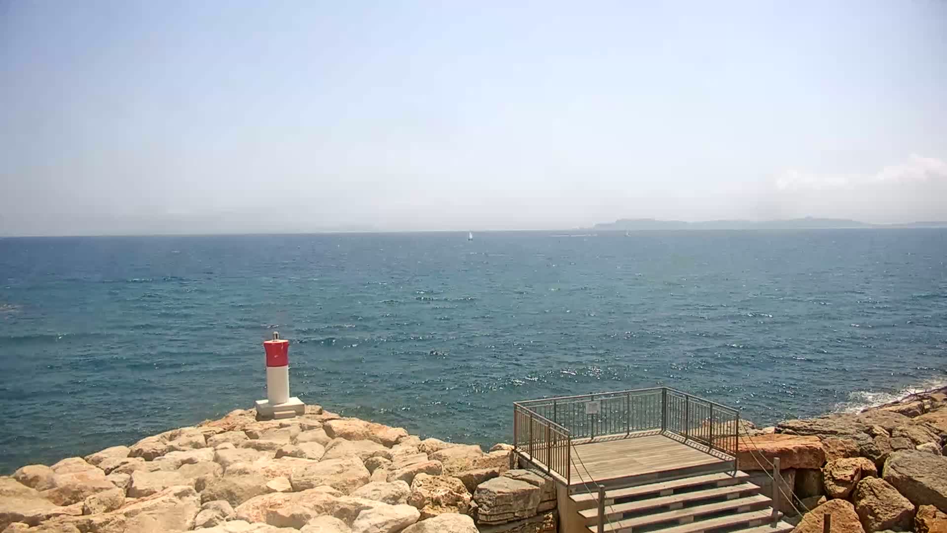 A small, red-and-white lighthouse stands on a rocky shoreline overlooking a calm, blue sea on a sunny day.