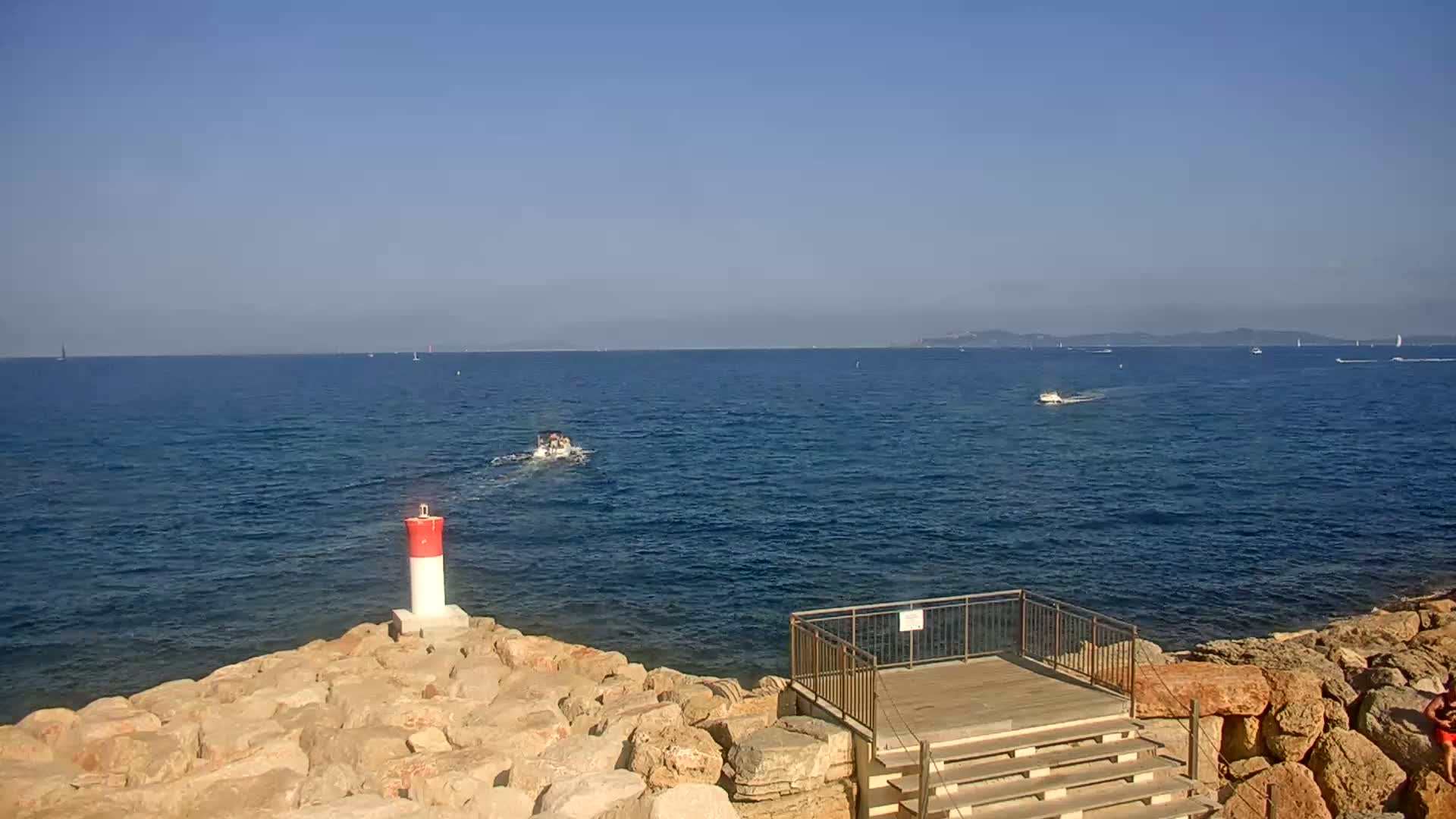 A rocky shoreline with a small lighthouse and a wooden viewing platform overlooks a calm, blue sea with several boats and a distant landmass under a clear blue sky.