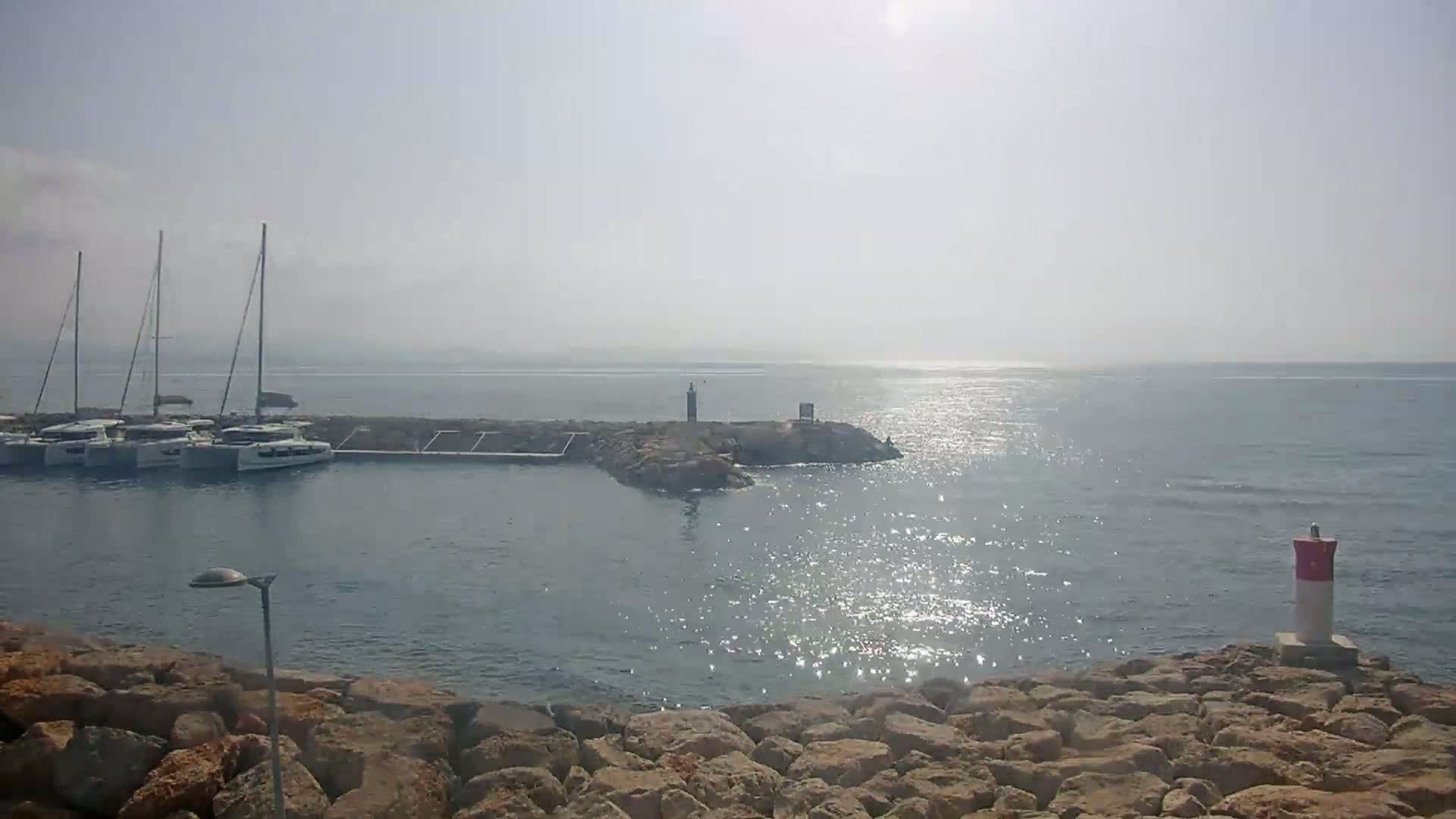 Several sailboats are moored in a calm, hazy harbor, partially sheltered by a rock jetty.