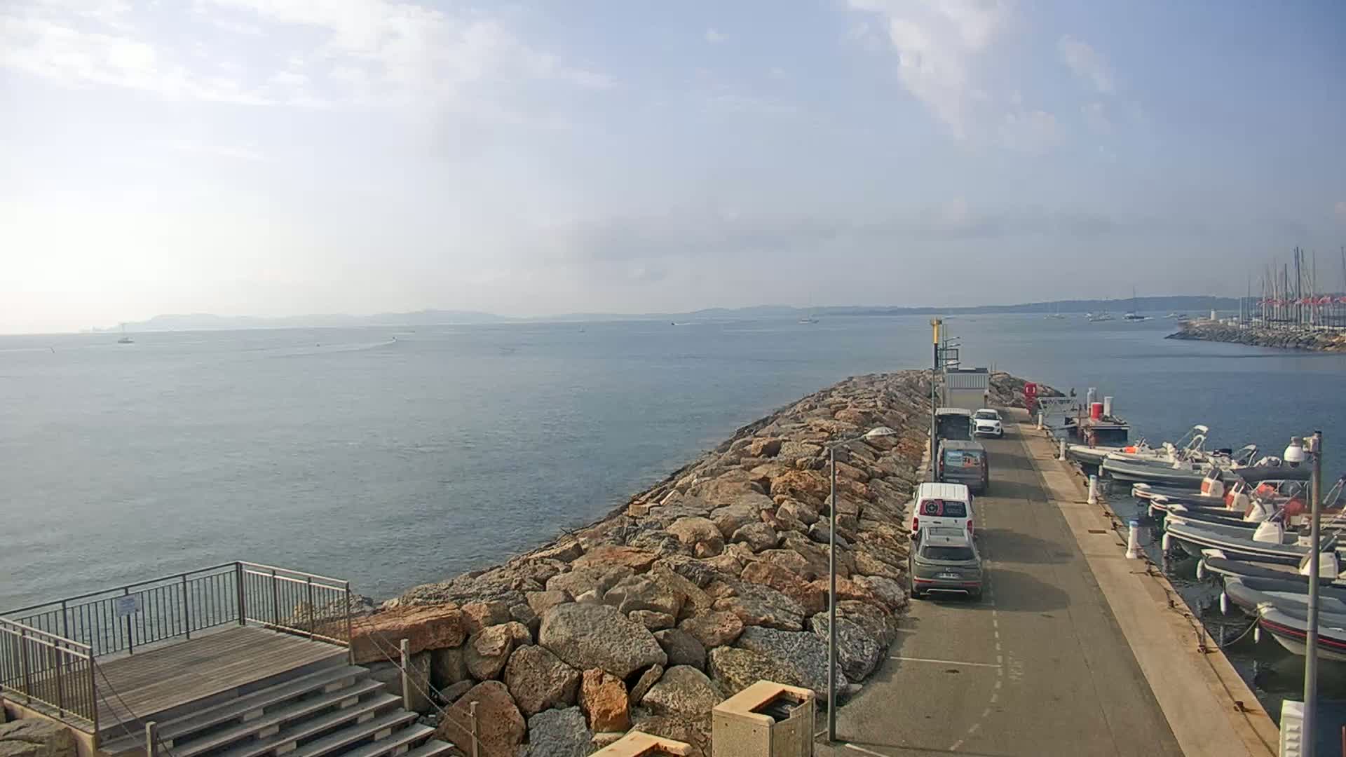 A rocky breakwater extends into a calm sea, with several vehicles parked along a paved road next to a small marina filled with boats under a mostly clear sky.