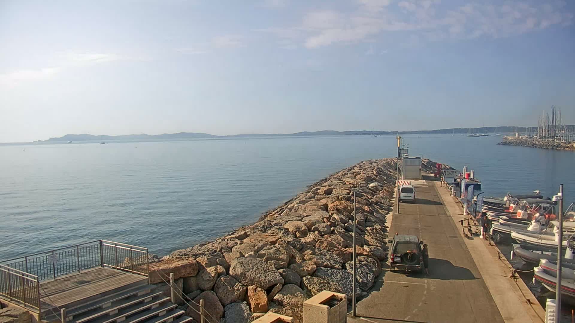 A rocky breakwater extends into a calm, blue sea under a clear sky, with several boats moored at a small harbor in the background.