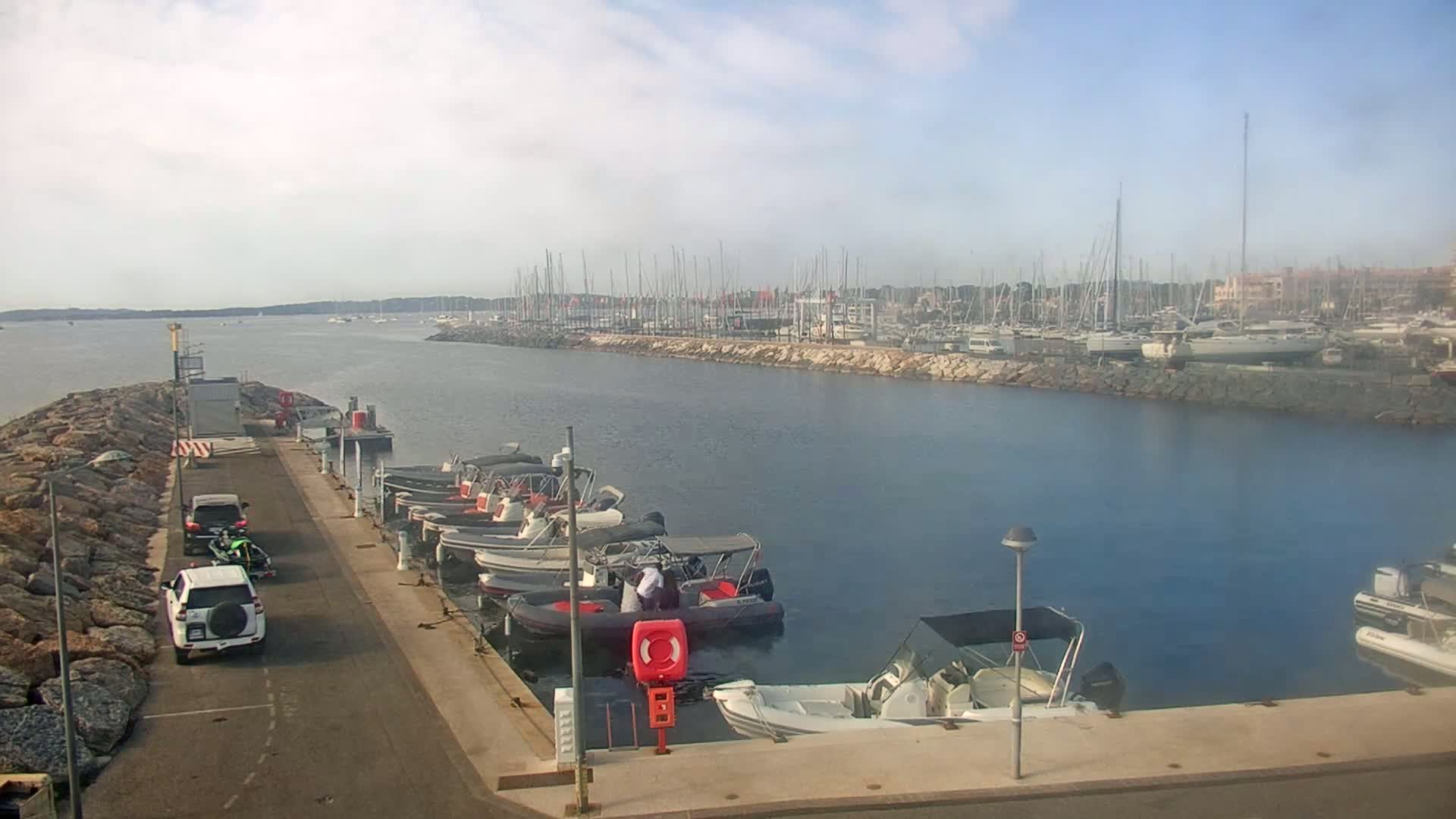 A marina with numerous boats docked, two vehicles parked nearby, and a calm body of water under a partly cloudy sky.