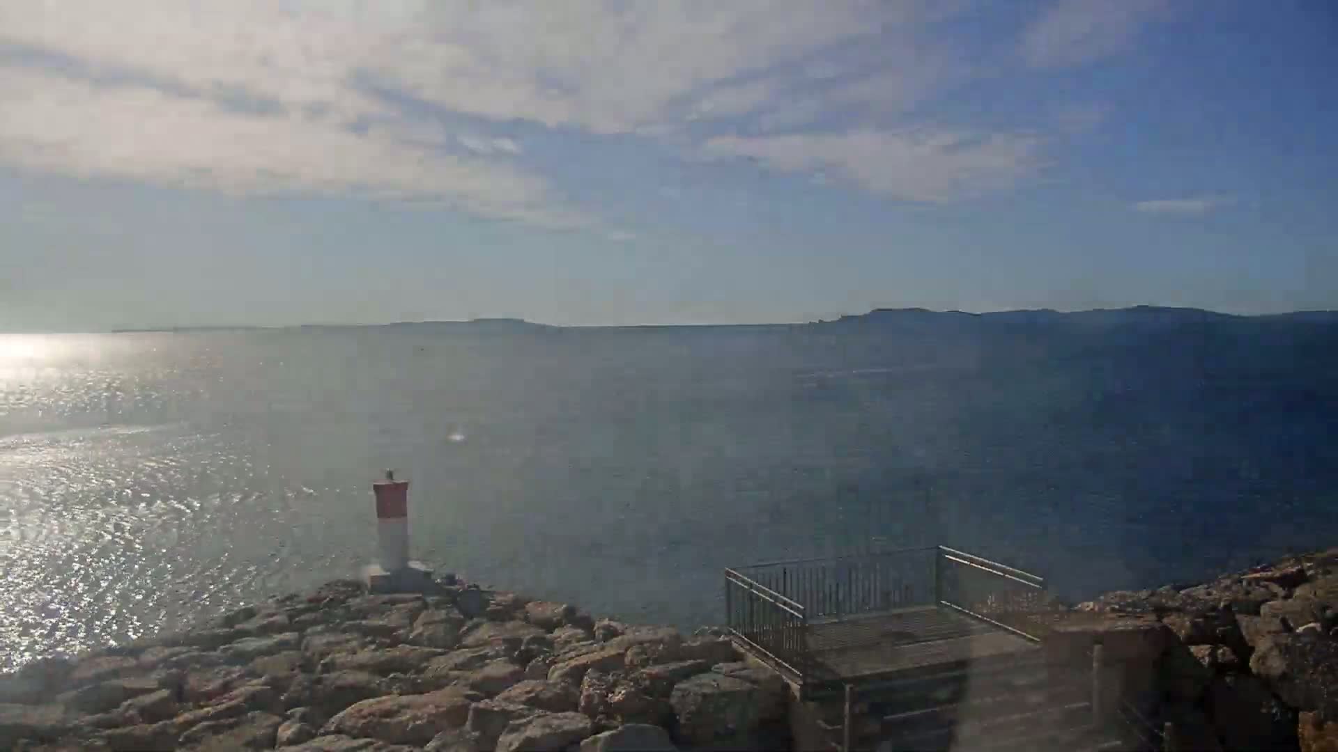 A rocky outcrop overlooks a calm, sunlit sea with distant landmasses under a partly cloudy sky.