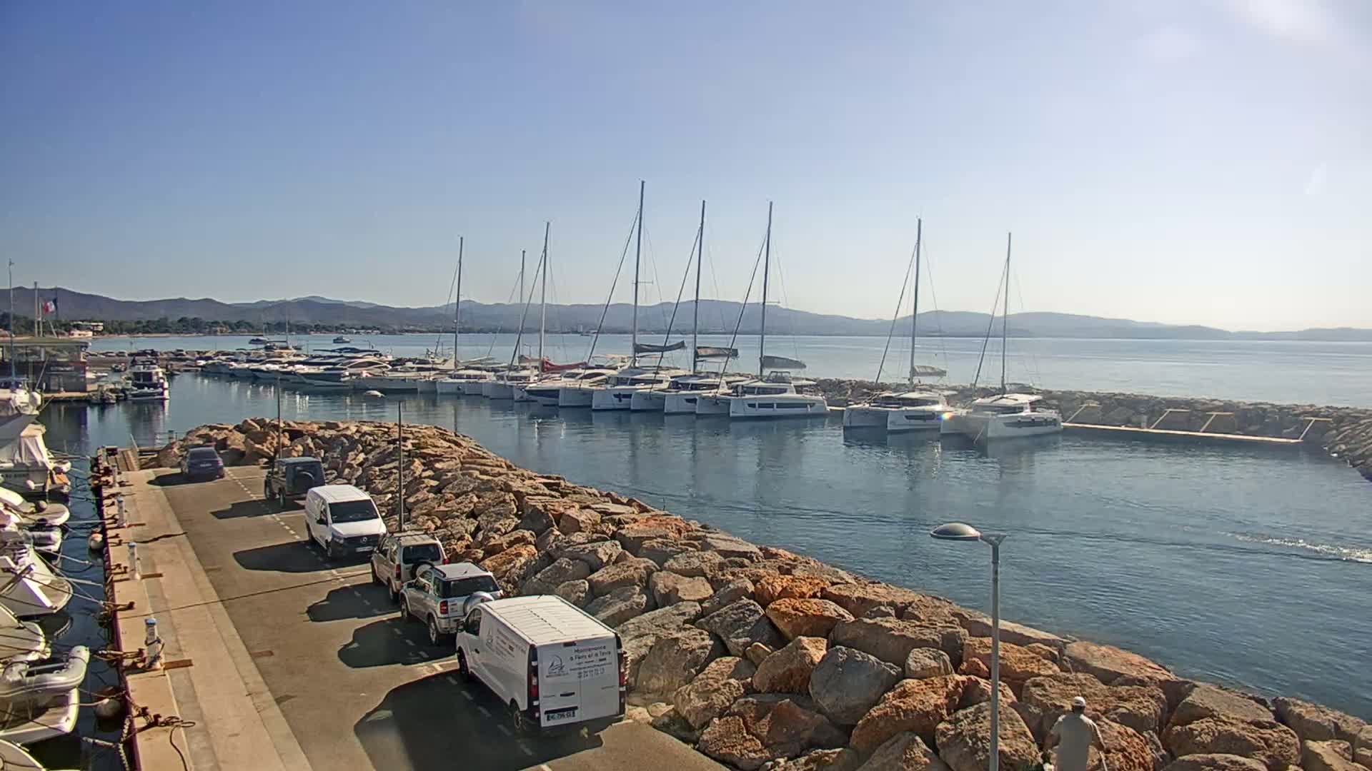 A marina filled with numerous sailboats and catamarans is nestled in a calm bay under a clear, sunny sky, with a roadway and several vehicles parked nearby.