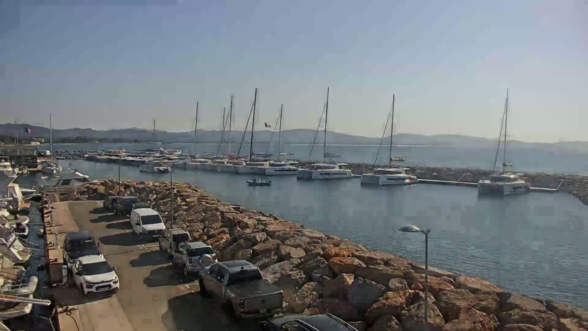 Numerous sailboats and yachts are docked in a marina on a sunny day, with cars parked along a rocky shoreline.