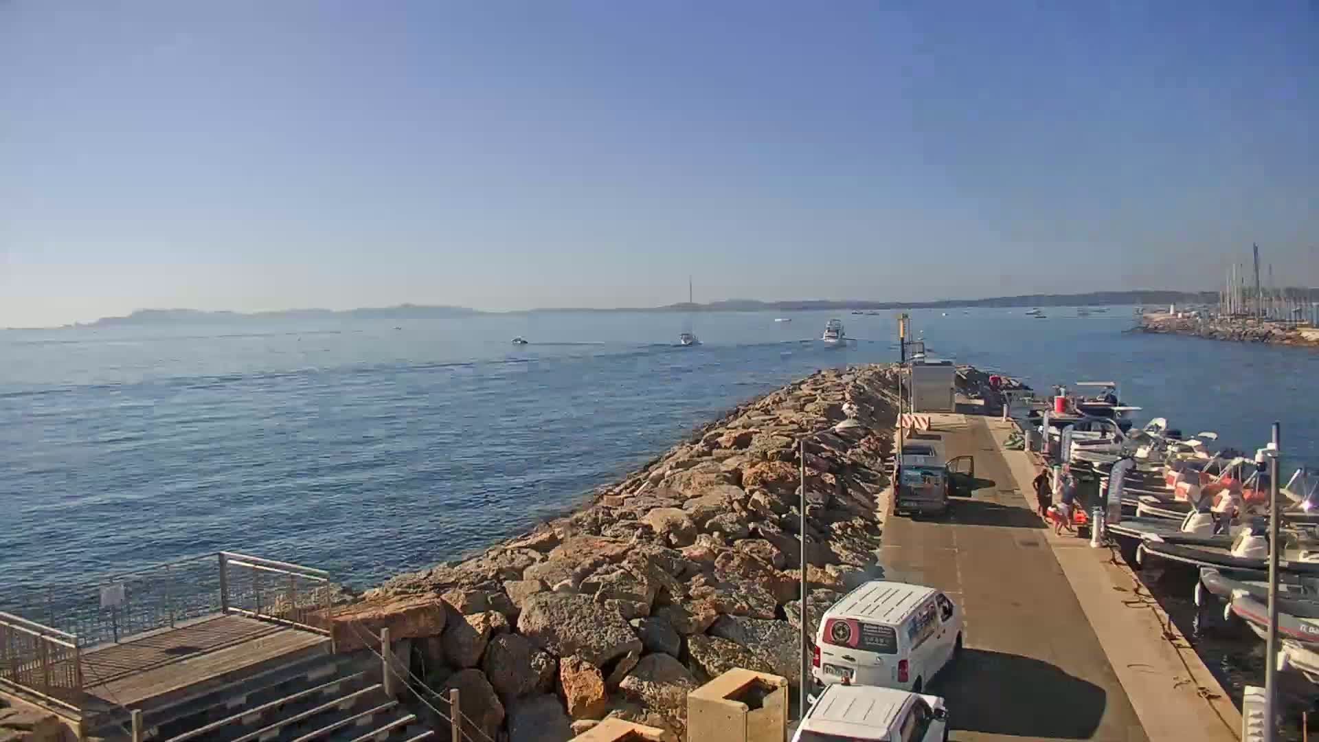 A rocky breakwater separates a calm, blue harbor with several boats moored from a clear, blue ocean with distant hills under a sunny sky.
