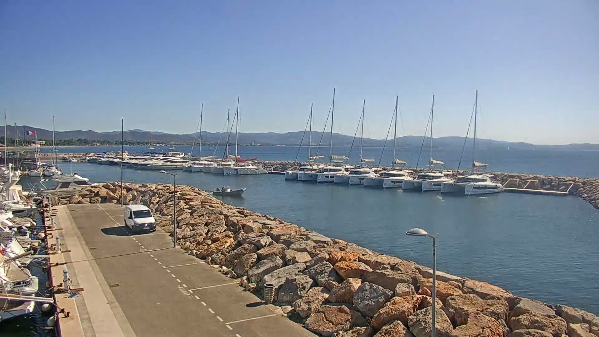A marina filled with sailboats and motorboats on a sunny day, is partially enclosed by a rocky wall with a van parked nearby.