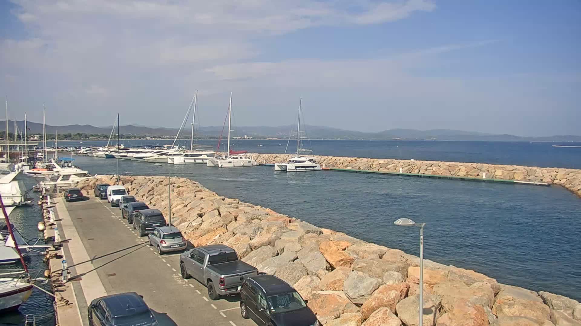 A marina filled with sailboats and motorboats on a sunny day, is partially enclosed by a rocky wall with a van parked nearby.
