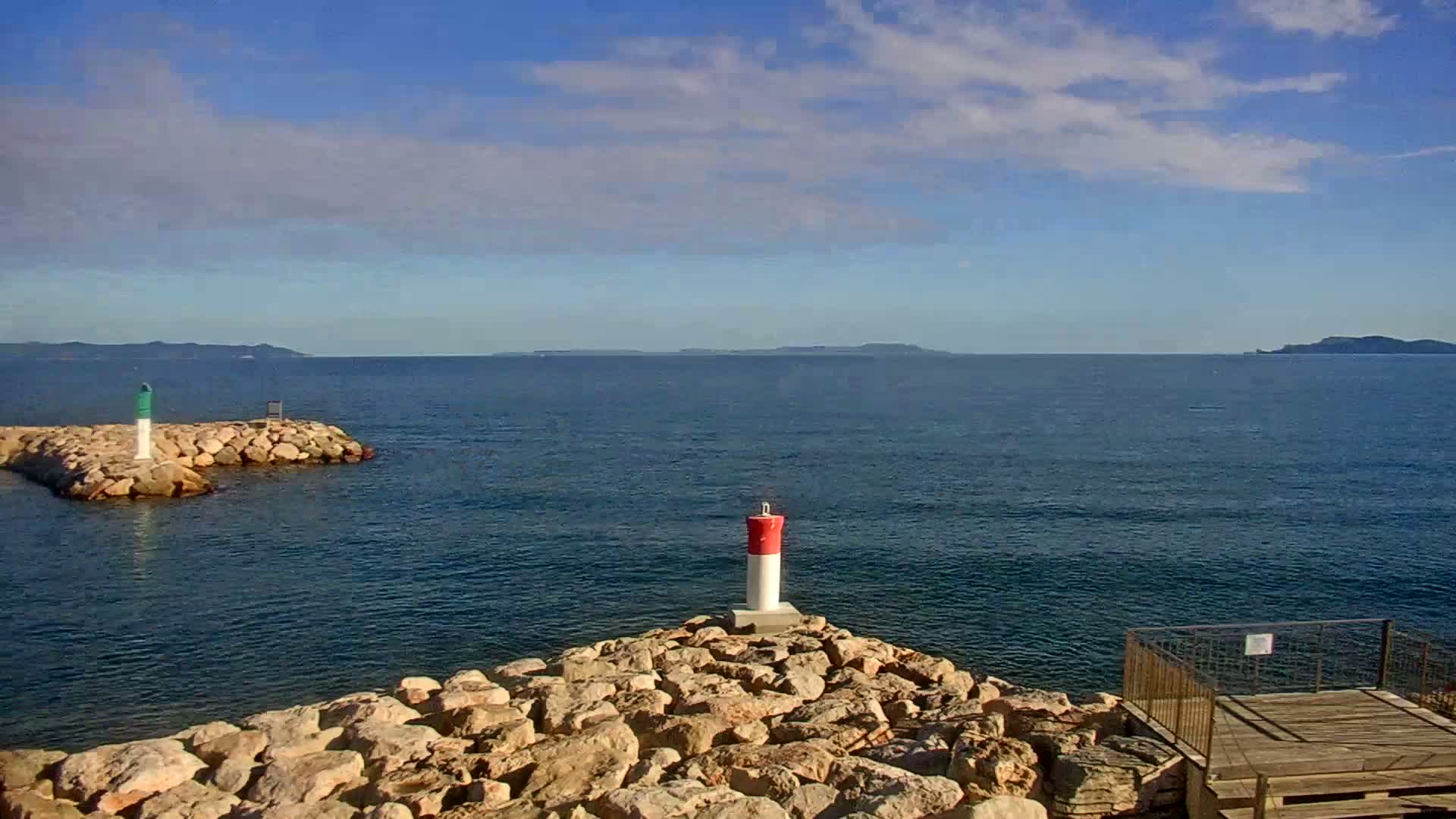 A marina filled with sailboats and motorboats on a sunny day, is partially enclosed by a rocky wall with a van parked nearby.