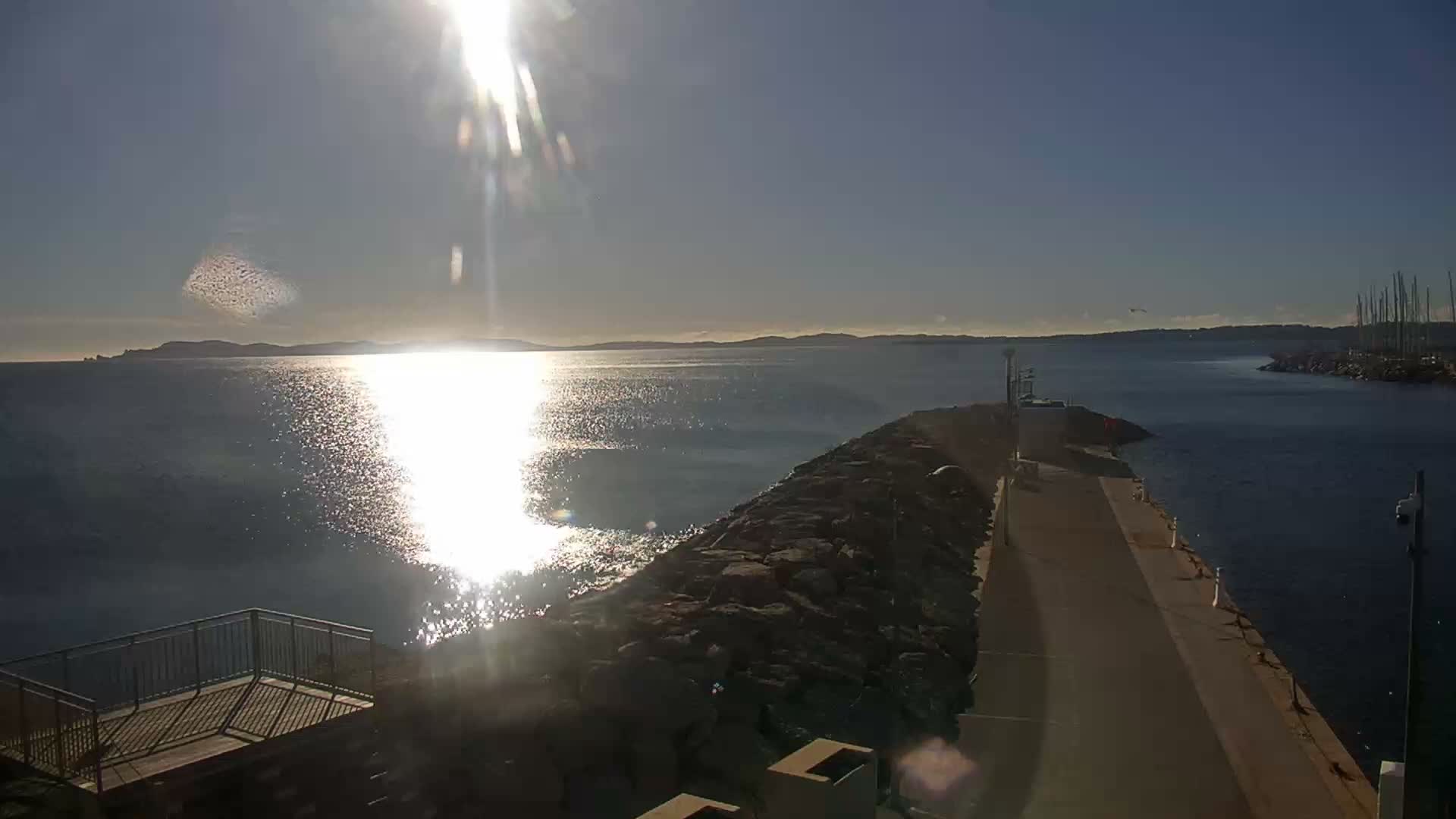 On a bright, clear day, the sun glares intensely across a calm expanse of ocean, bordered by a rocky breakwater and a pier that extends towards a distant coastline and a marina filled with boat masts.