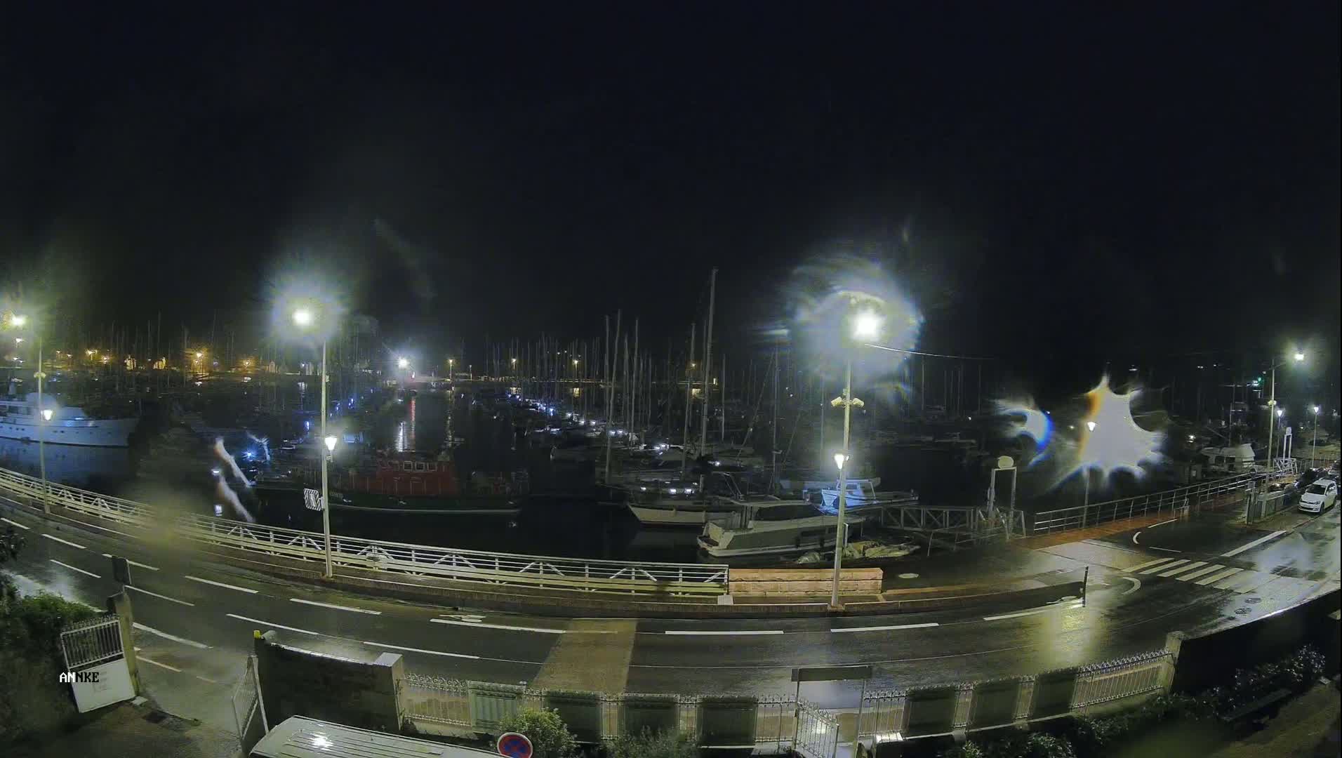 A nighttime view of a harbor filled with boats under a dark, possibly stormy sky, with rain on the wet road next to the water.