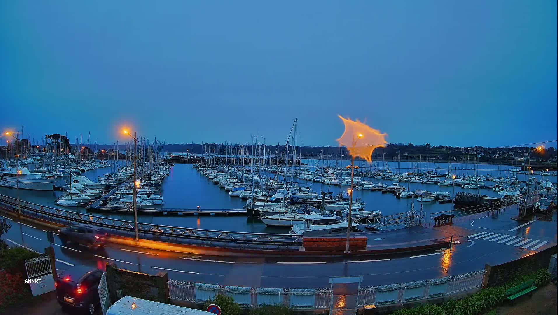 A marina filled with numerous sailboats and motorboats is seen at dusk under a dark blue sky, with rain on the wet roads nearby.