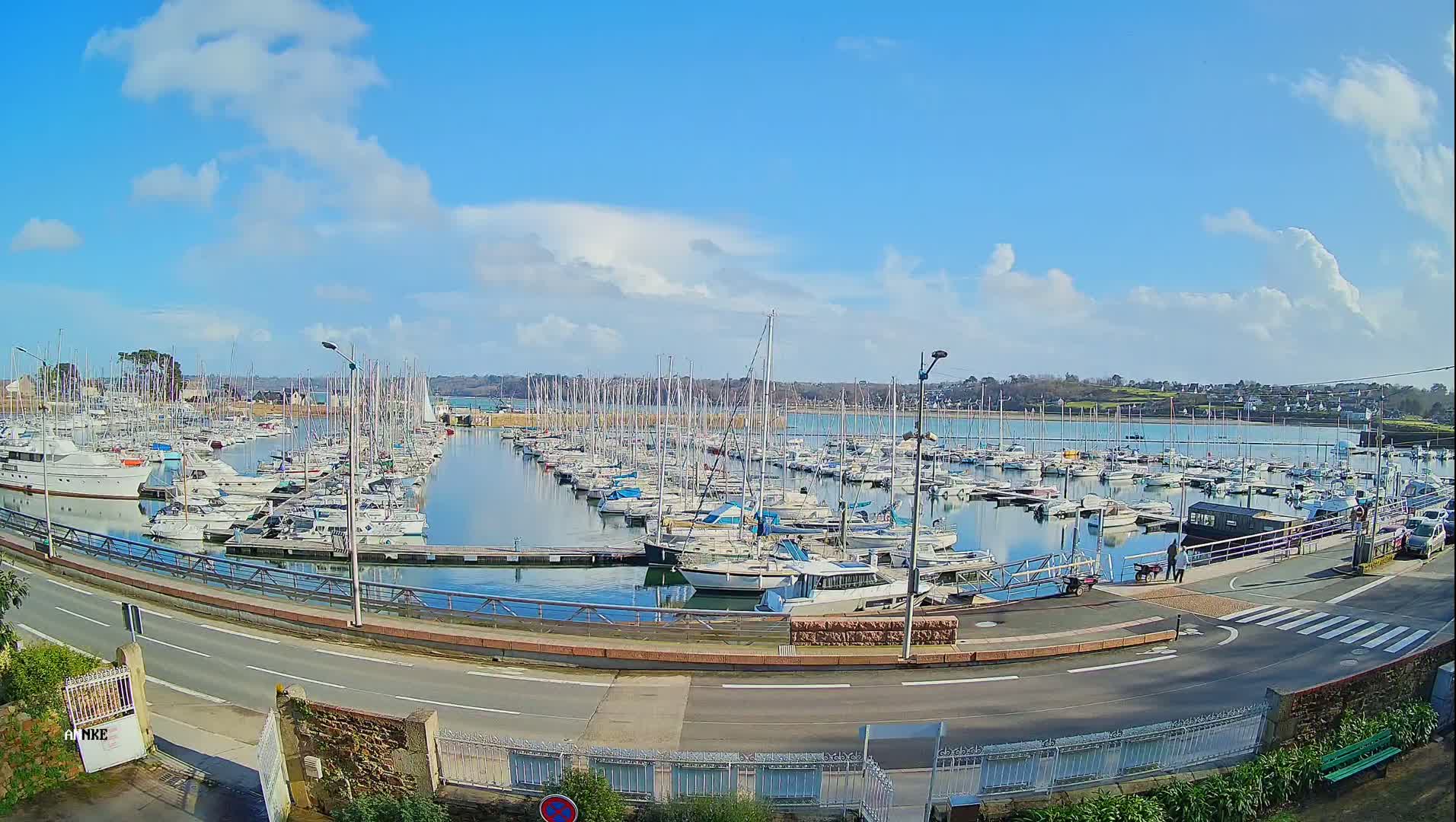 A marina filled with numerous sailboats and motorboats is situated on calm water under a mostly sunny sky with scattered clouds.