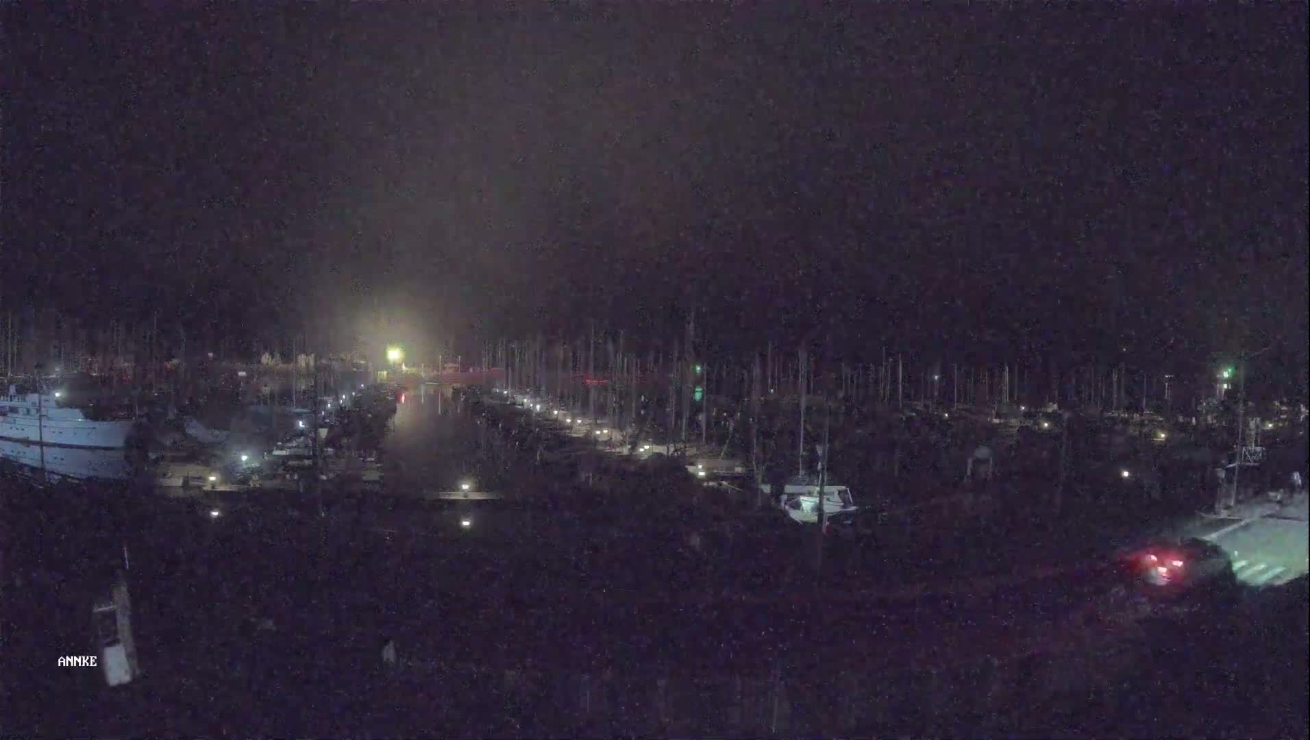 A nighttime aerial view shows a marina filled with boats, lit by several small lights along a dock, under a dark, cloudy sky.