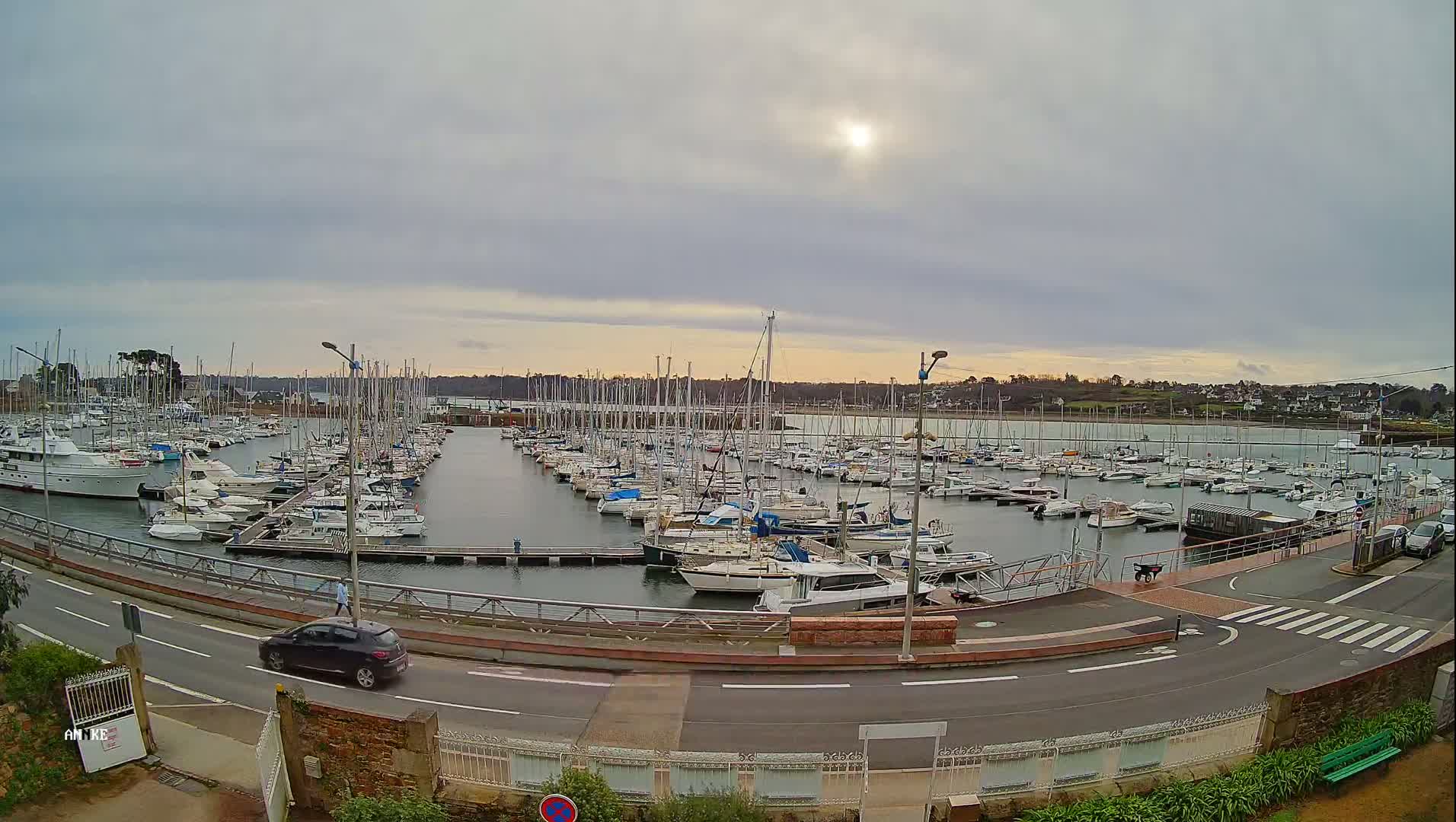 A marina filled with numerous sailboats and motorboats is seen under an overcast sky, with a road and a few cars visible in the foreground.