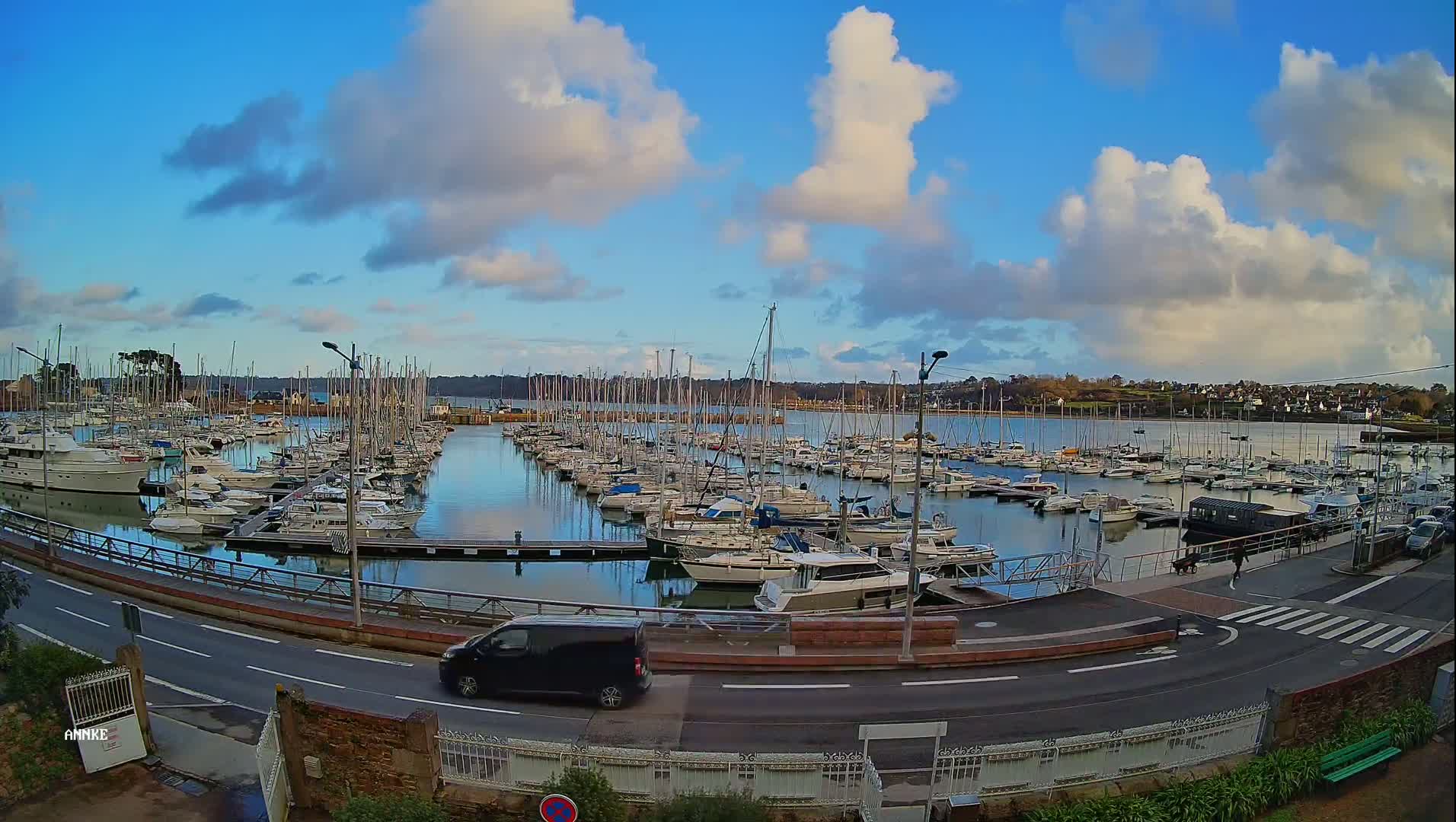 A marina filled with numerous sailboats and motorboats is seen under a partly cloudy sky, with a road and a vehicle visible in the foreground.