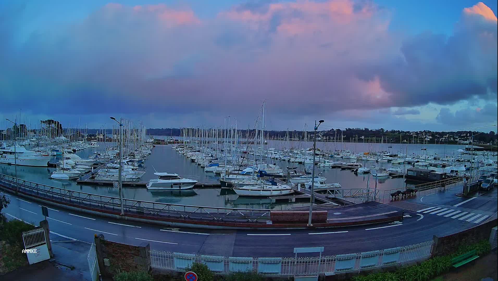 A marina filled with numerous sailboats and motorboats is shown under a cloudy sky with pink hues near the horizon.